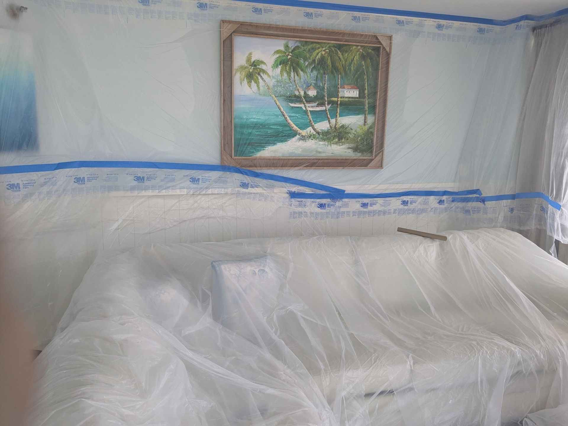 Room prepared for painting; walls, sofa, and art covered in plastic sheeting and painter's tape.