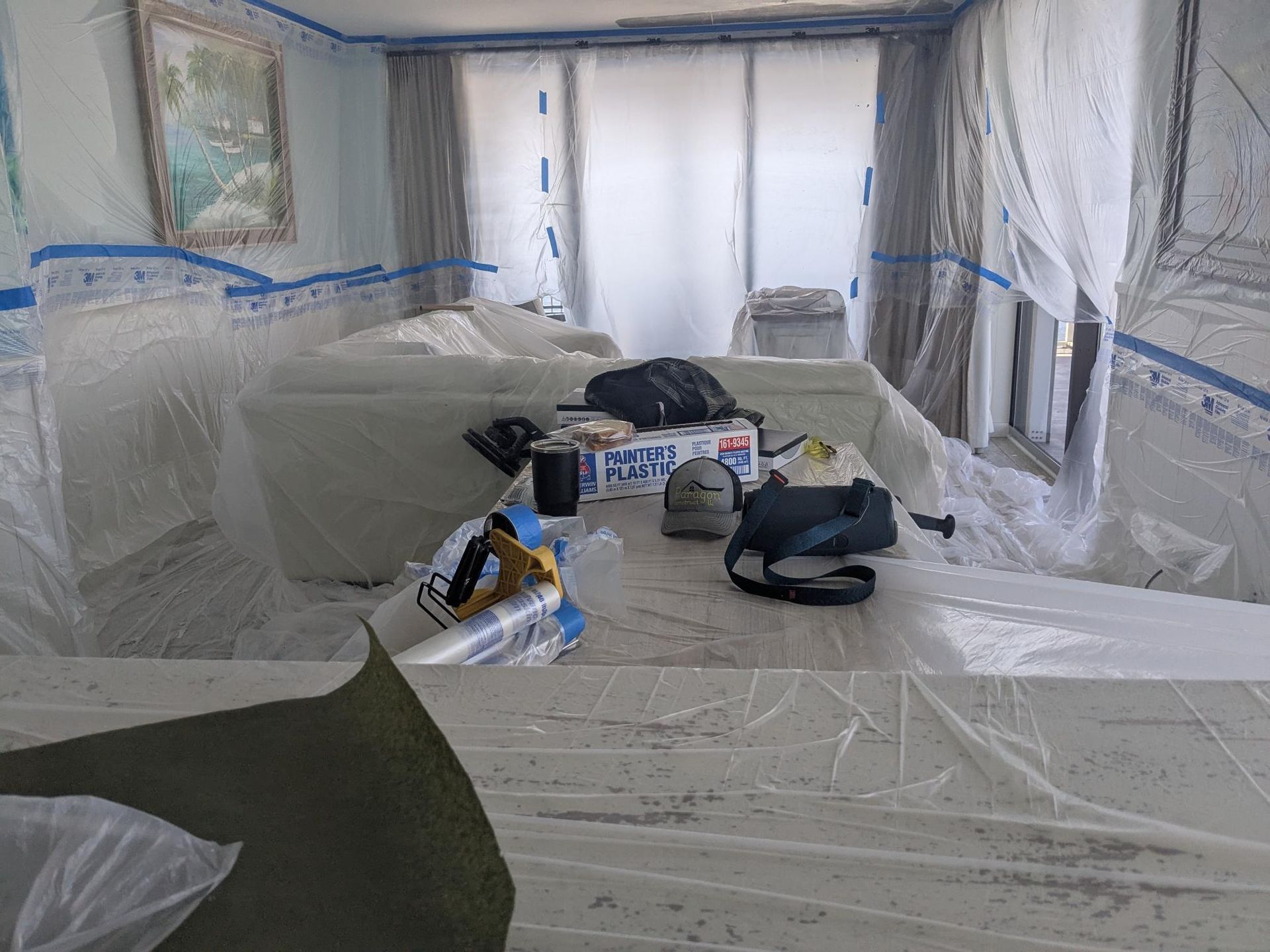 Room covered in plastic sheeting for painting; tools and items on the sofa and floor.