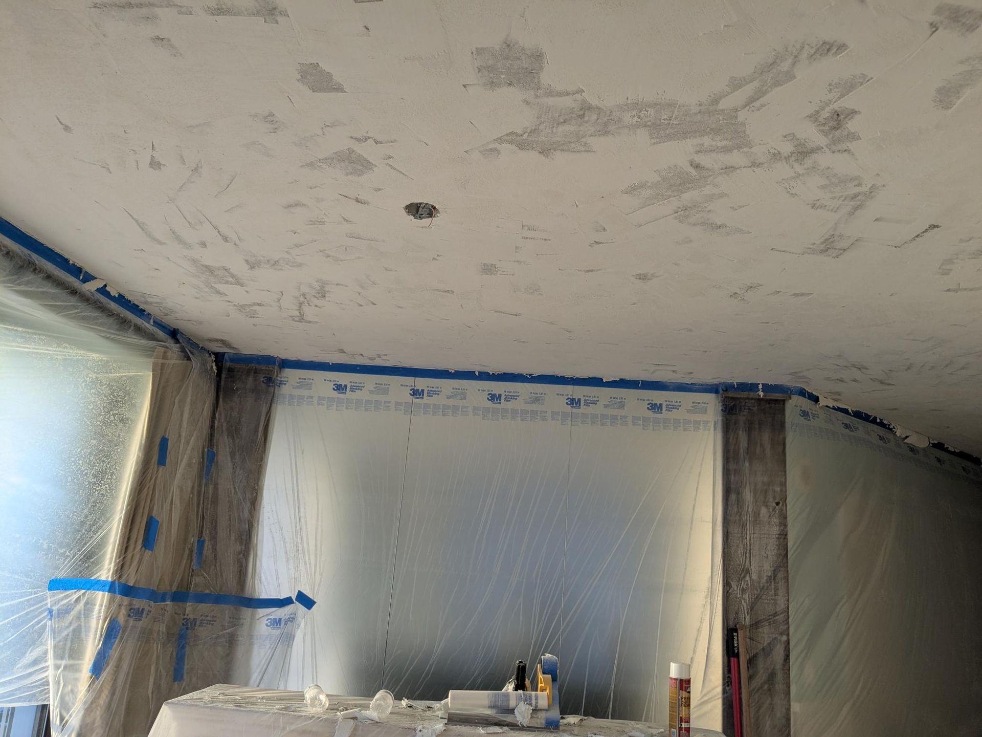 Interior room under renovation, with taped windows and textured ceiling. Blue tape lines the edges.