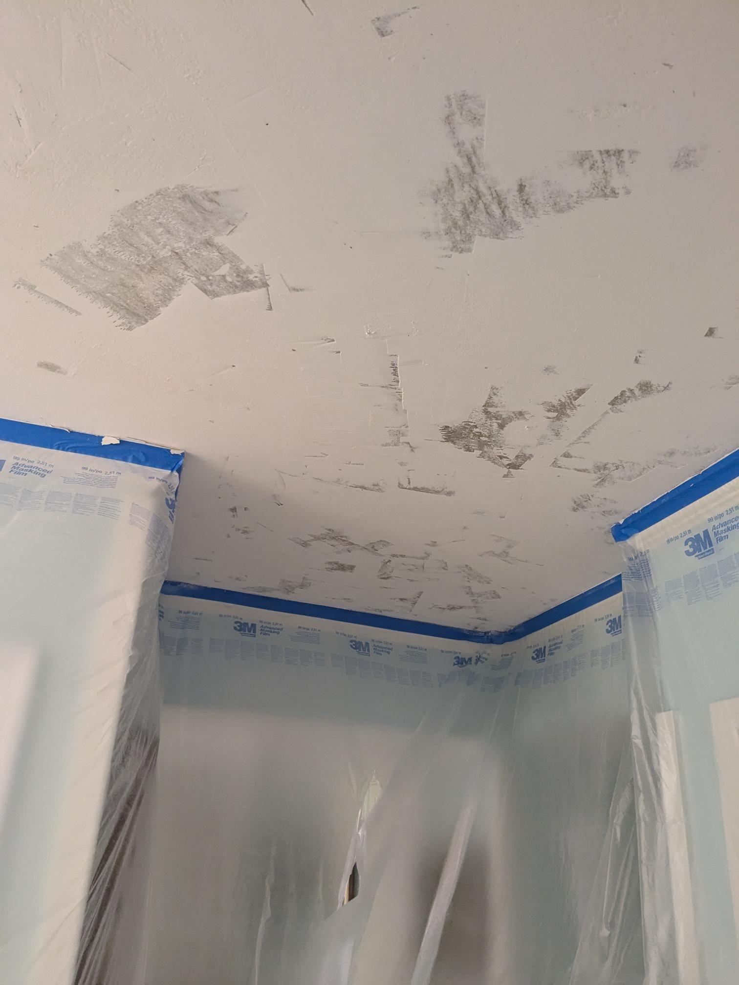 Ceiling with peeling paint, corners taped with blue painter's tape, plastic sheeting covers walls.