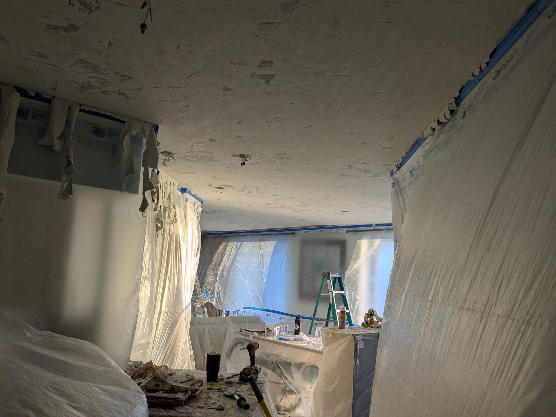 Interior room undergoing renovation, covered in plastic sheeting. Blue tape marks areas.