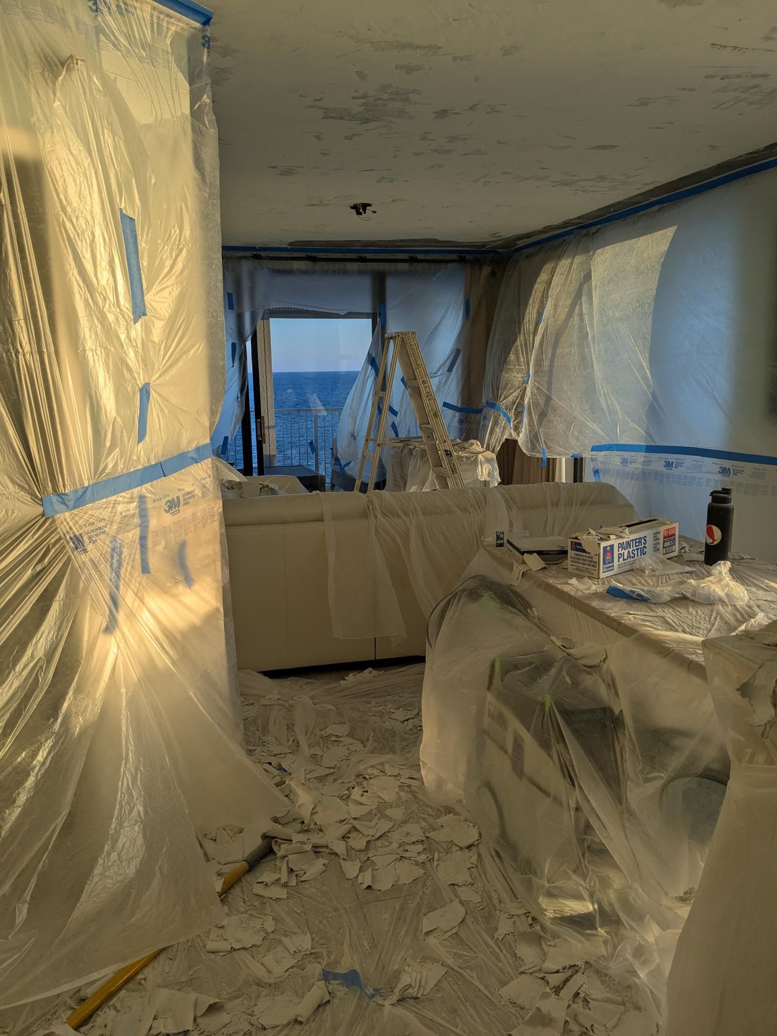 Room under renovation, covered in plastic sheeting. Debris litters the floor; a ladder leans against the window.