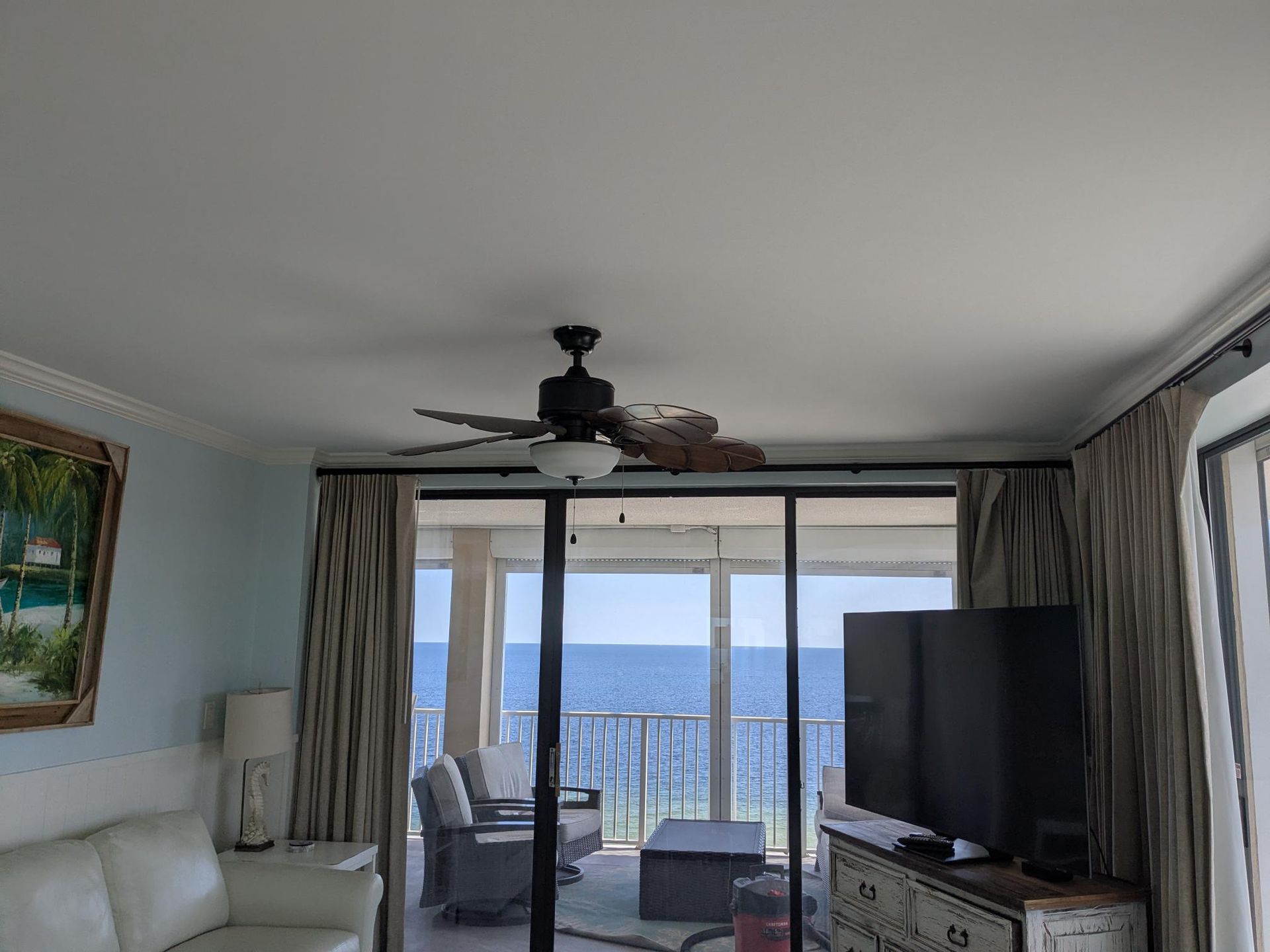 Living room with ocean view, sliding glass doors, ceiling fan, TV, sofa, and curtains.