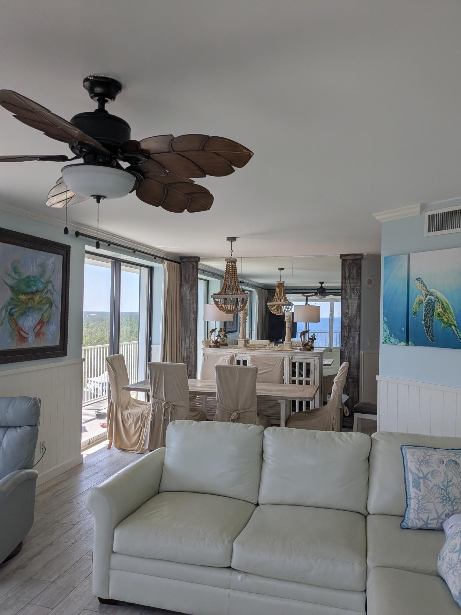 Coastal-themed living room with ocean view. Features a white sofa, dining table, art, and ceiling fan.