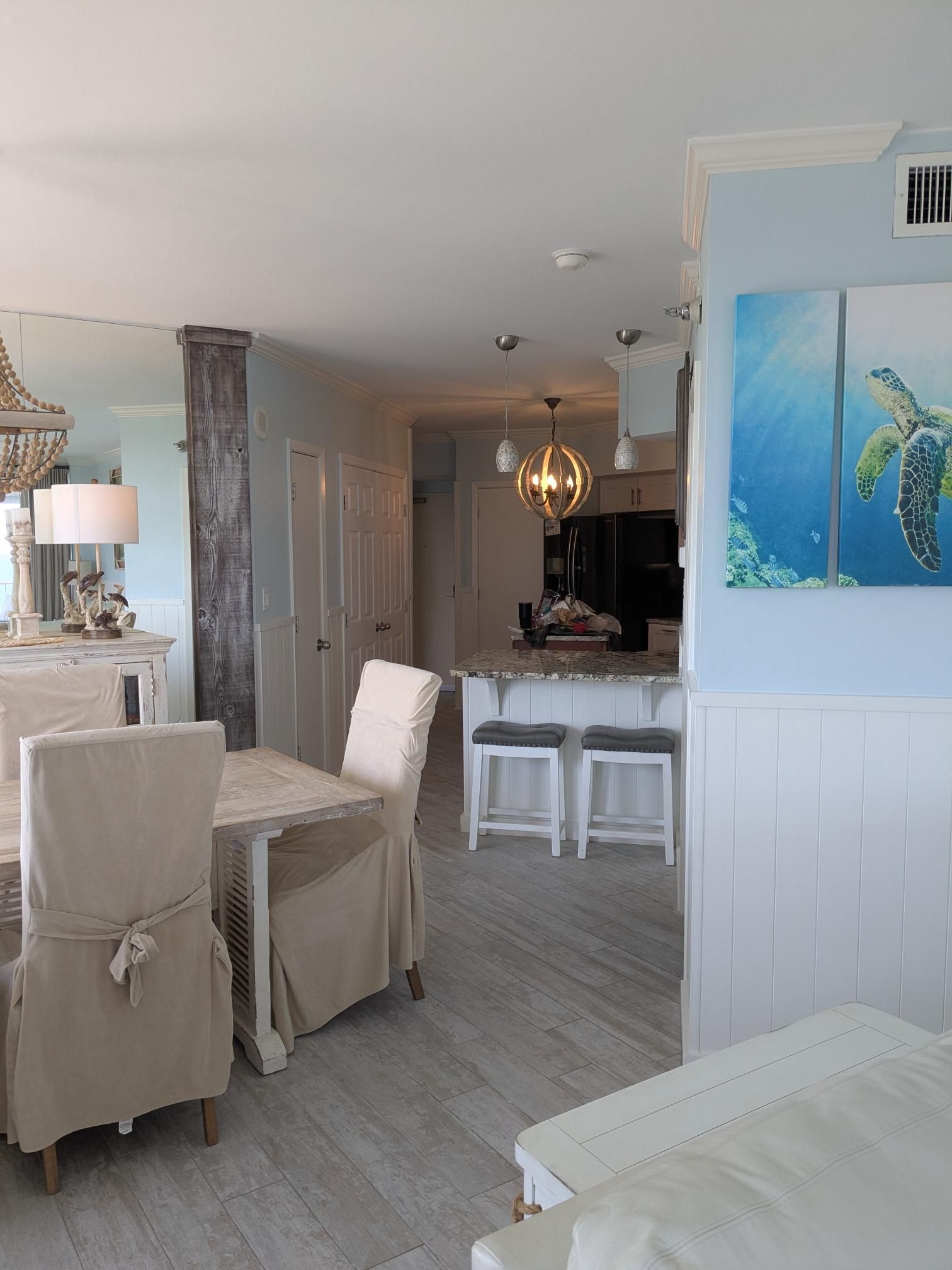 Coastal-themed dining room: dining table with slipcovered chairs, kitchen with island and bar stools, and sea turtle art.