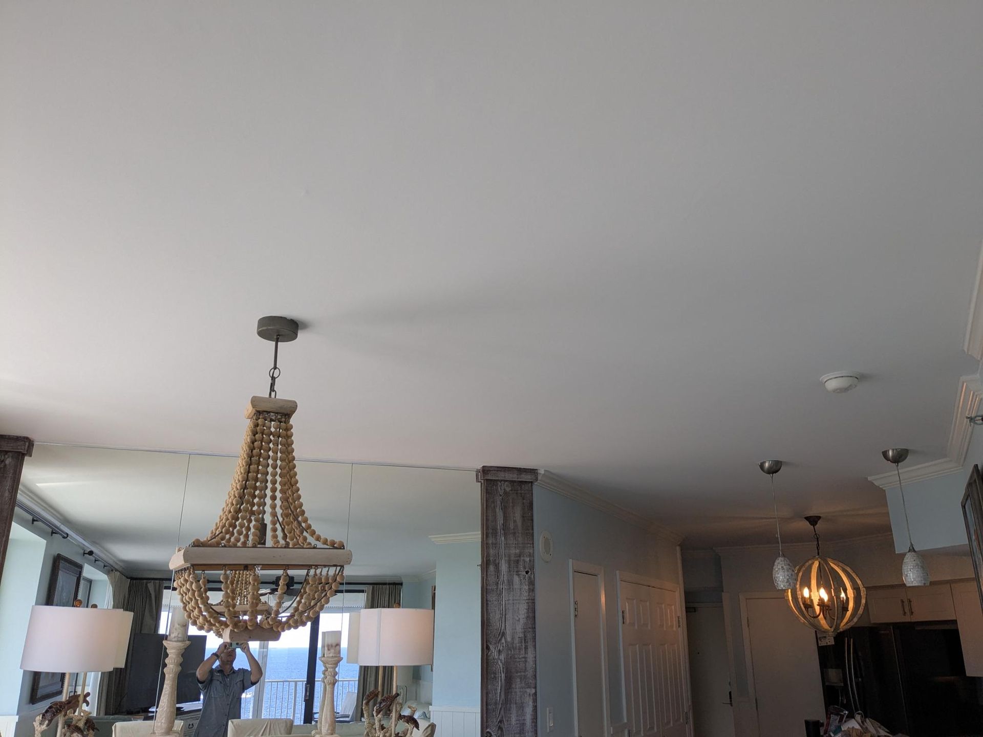 Wooden beaded chandelier hanging from a white ceiling, with other light fixtures and a room in the background.