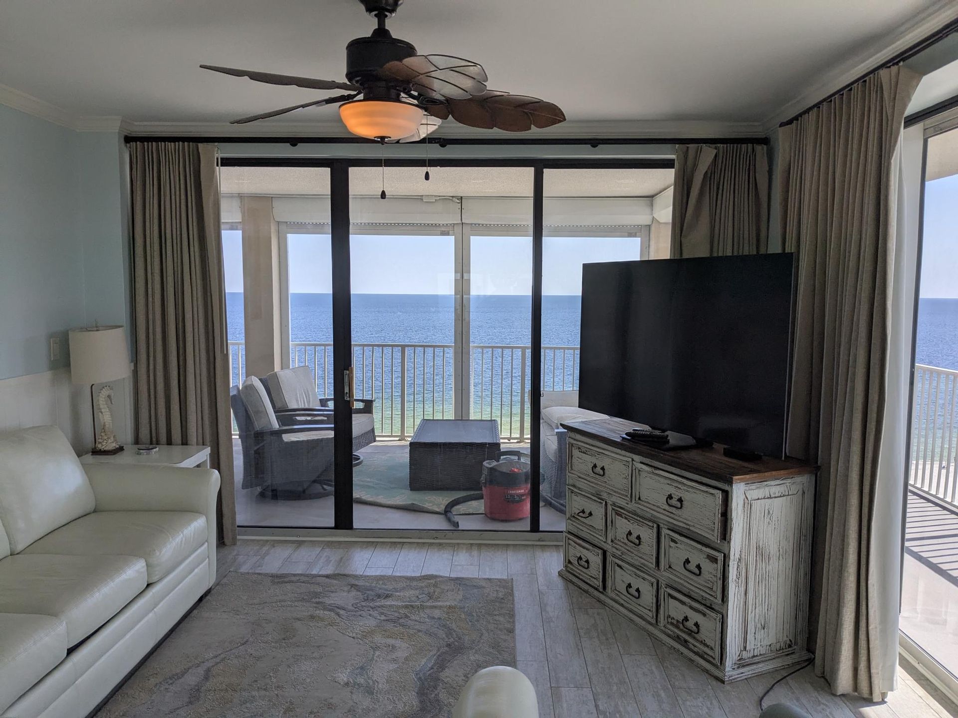 Living room with ocean view, white couch, TV on a distressed cabinet, sliding glass doors, and beige curtains.
