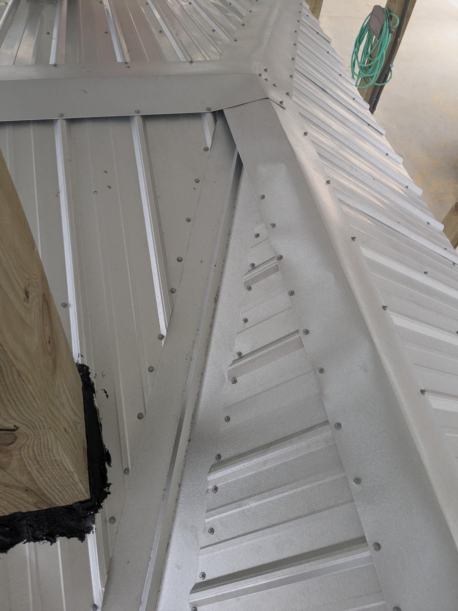 Close-up of a light gray metal roof corner being installed, with visible fasteners. A wooden beam is in the lower left.