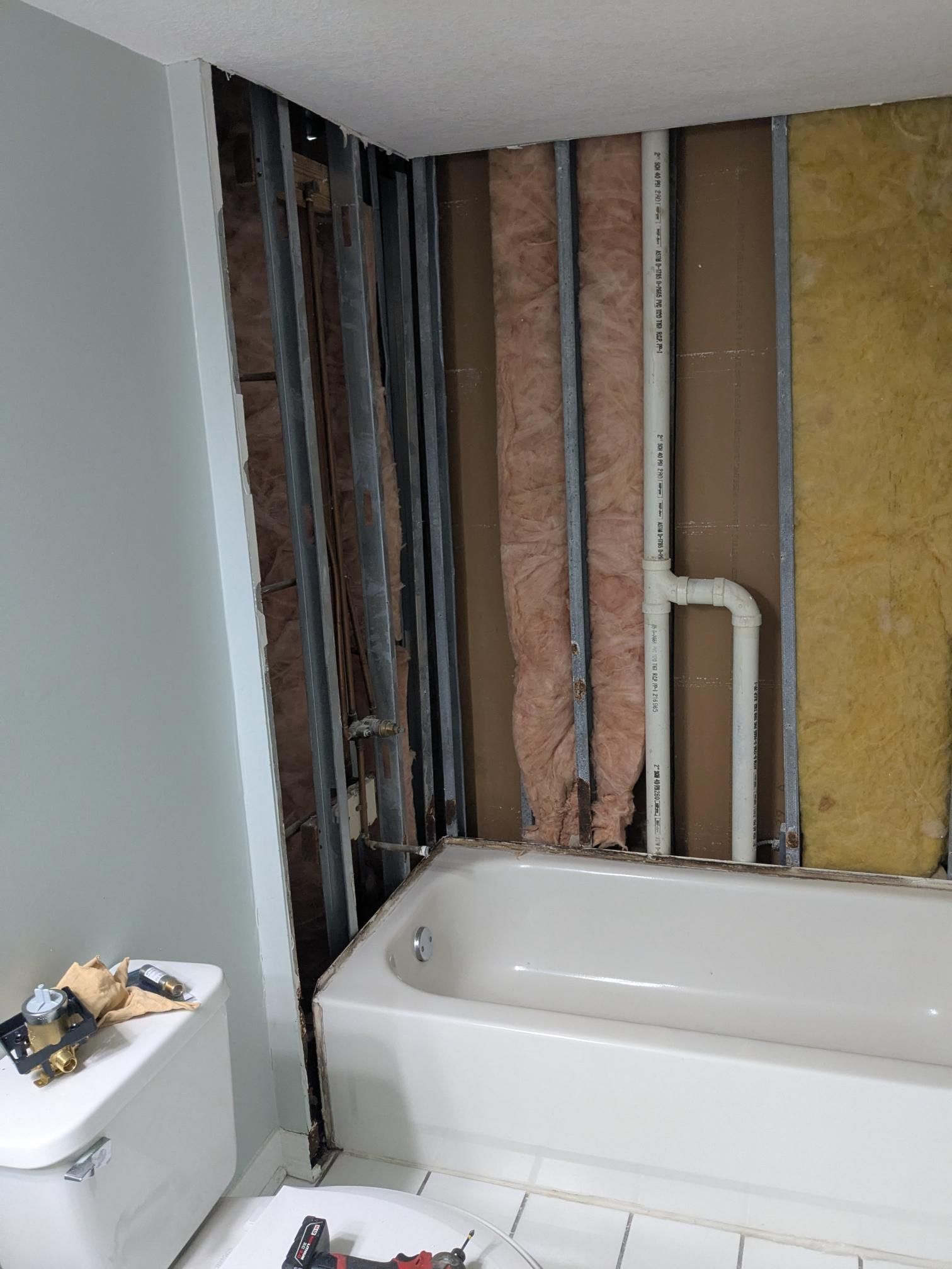 Bathroom wall under renovation, exposing studs, insulation, and plumbing near a bathtub and toilet.