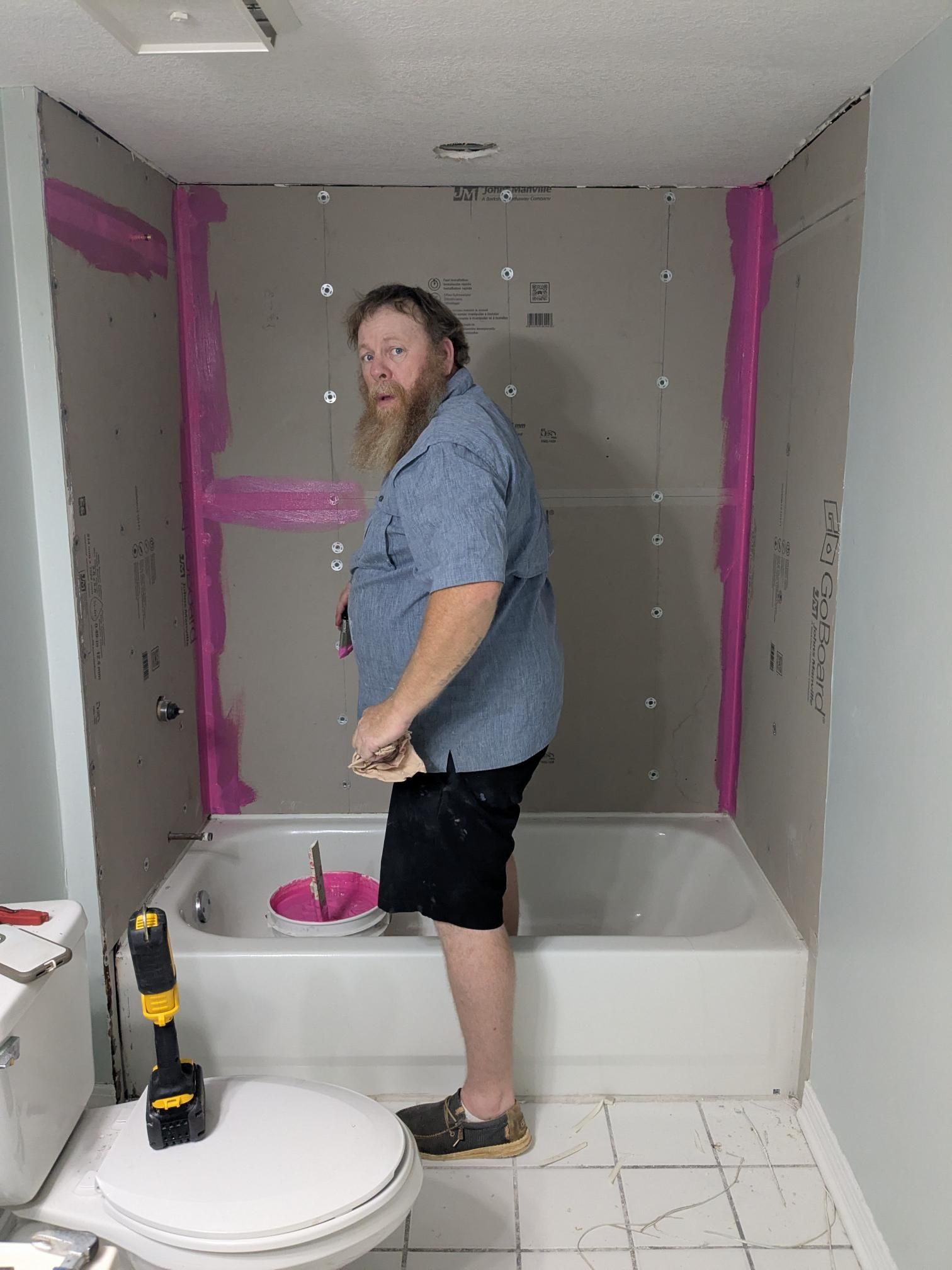 Man standing in a bathroom, near bathtub, working on the walls; pink sealant visible.