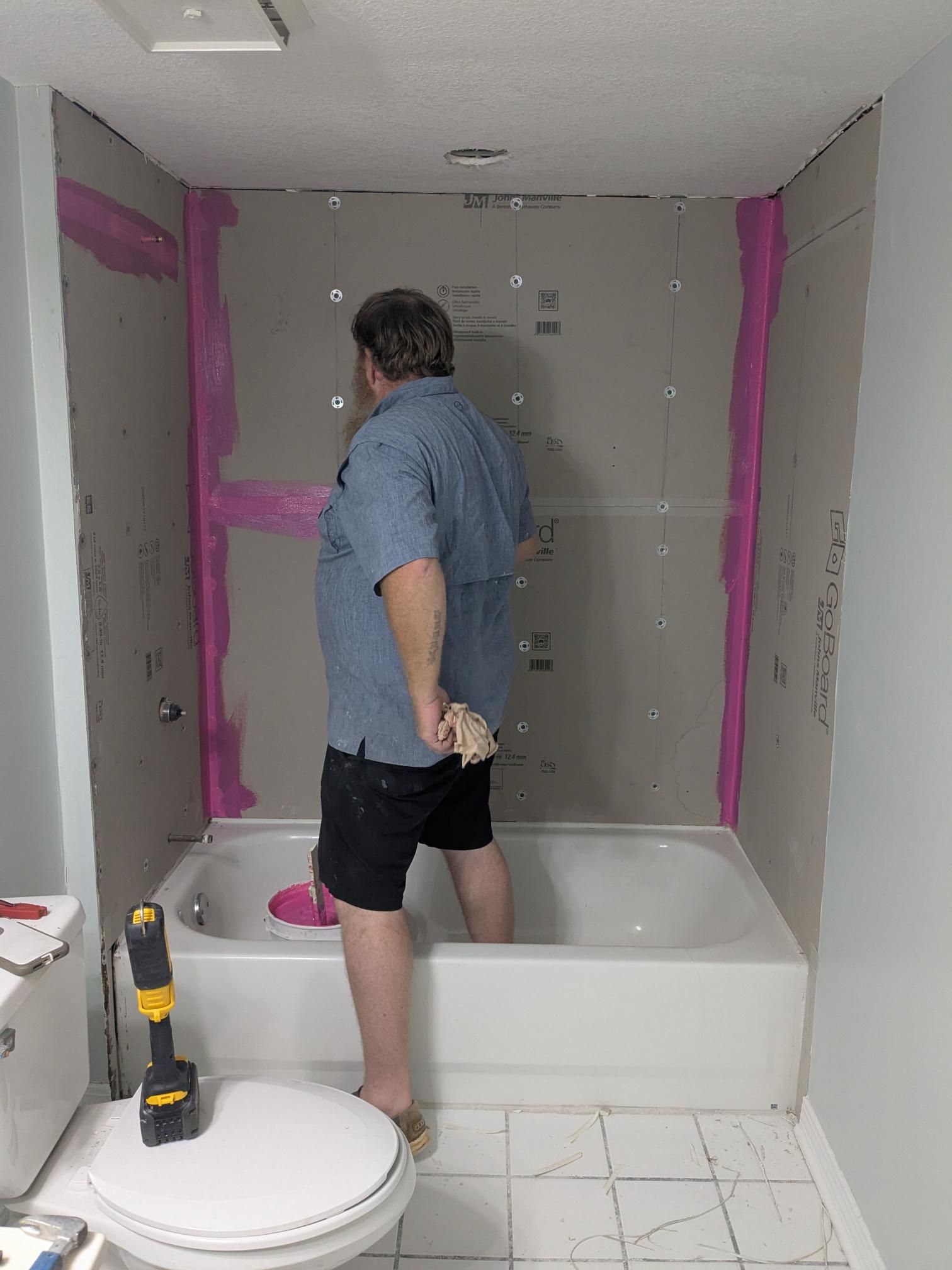 A person stands in a bathtub during bathroom renovation. Gray walls with pink sealant. Drill sits nearby.
