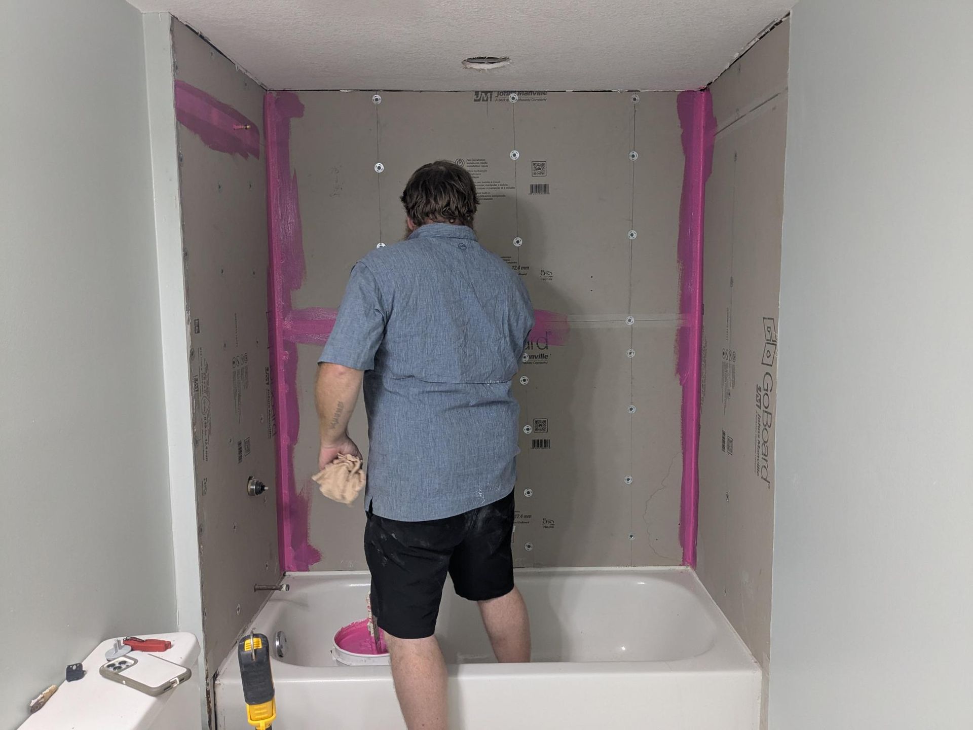 Person standing in a bathtub, wiping the wall. Pink sealant outlines the shower area. Light blue walls.
