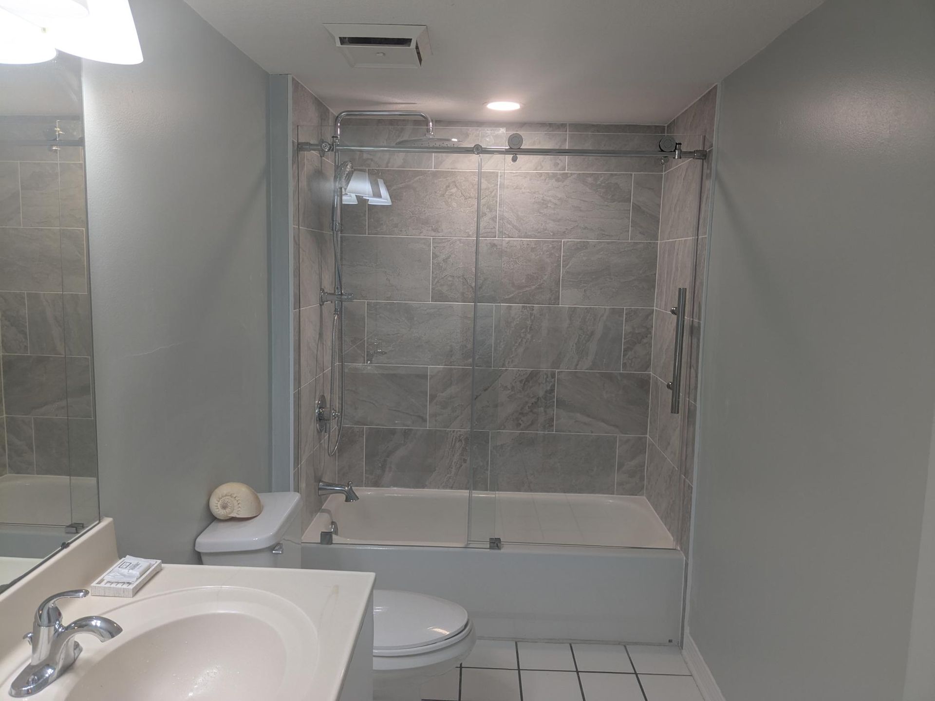 Bathroom with gray tiled shower and white tub, sink, and toilet. Gray walls.