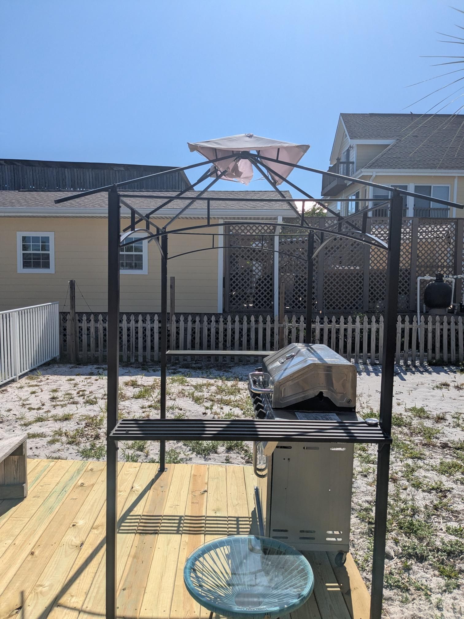 Gazebo over grill on a wooden deck in a sandy yard. Umbrella on top; two houses in background.