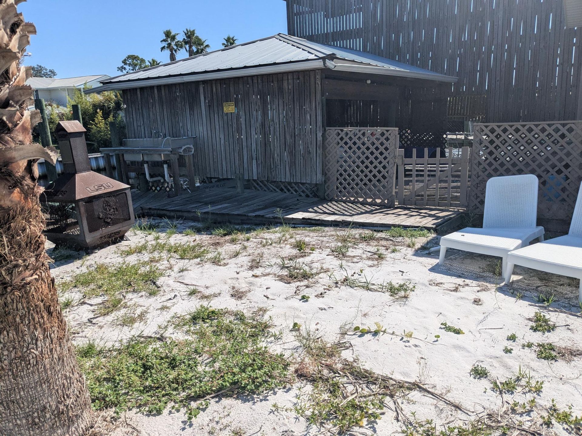 A backyard scene with a small tiki hut, two white chairs, and an old grill on a sandy surface.