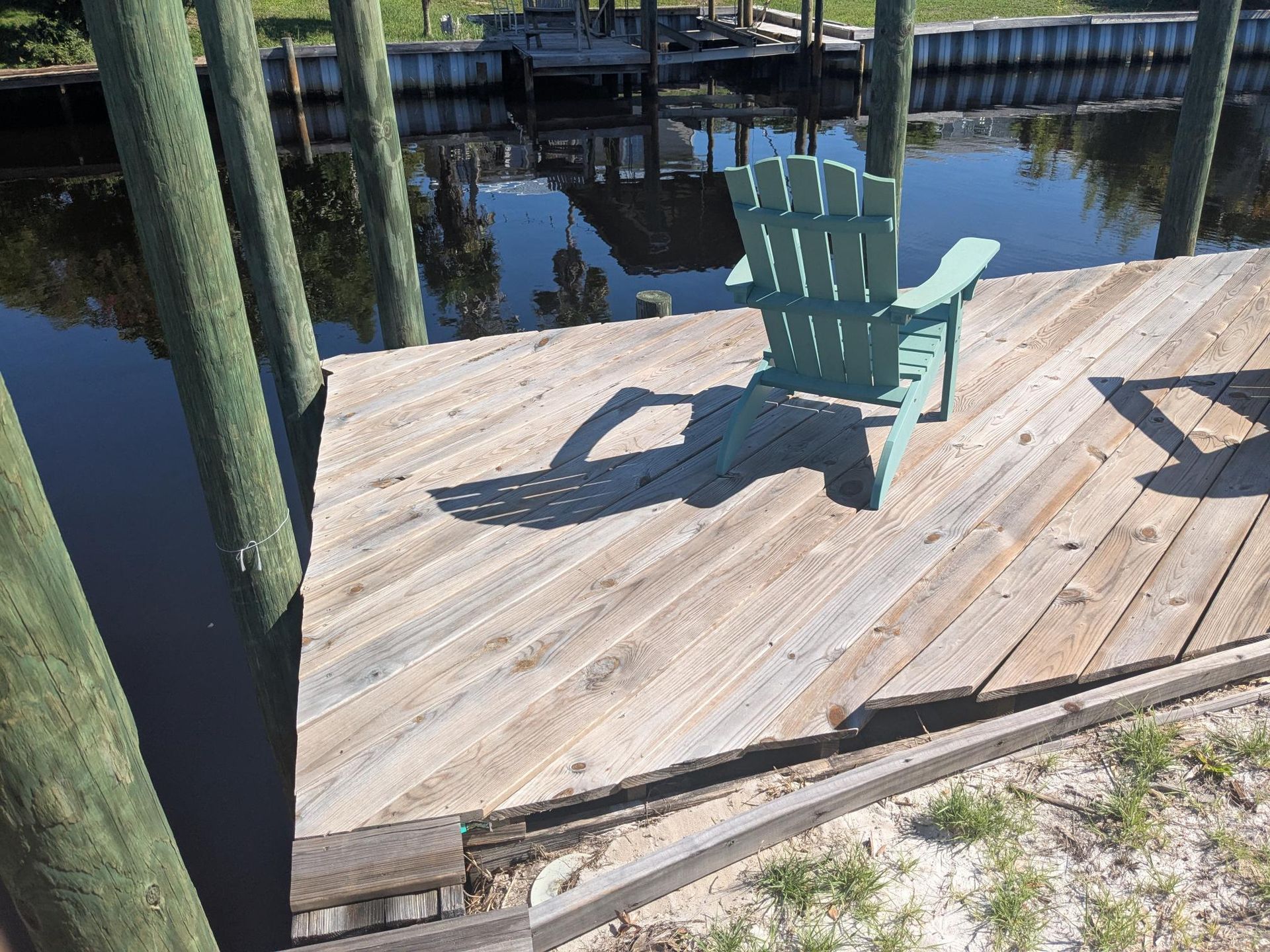Wooden dock with green Adirondack chair, overlooking water. Sunlight casts shadows.
