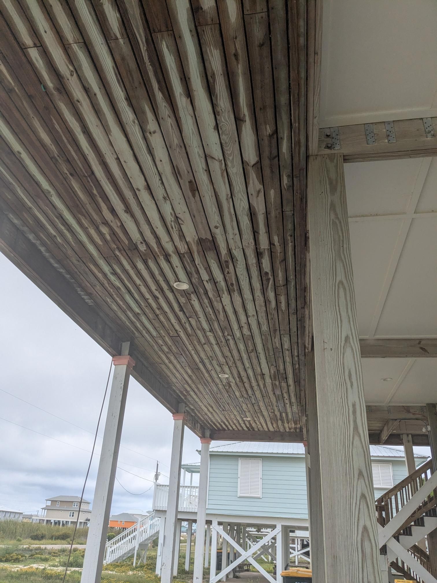 Wooden porch ceiling with weathered boards, supported by posts, overlooking a beach with buildings.