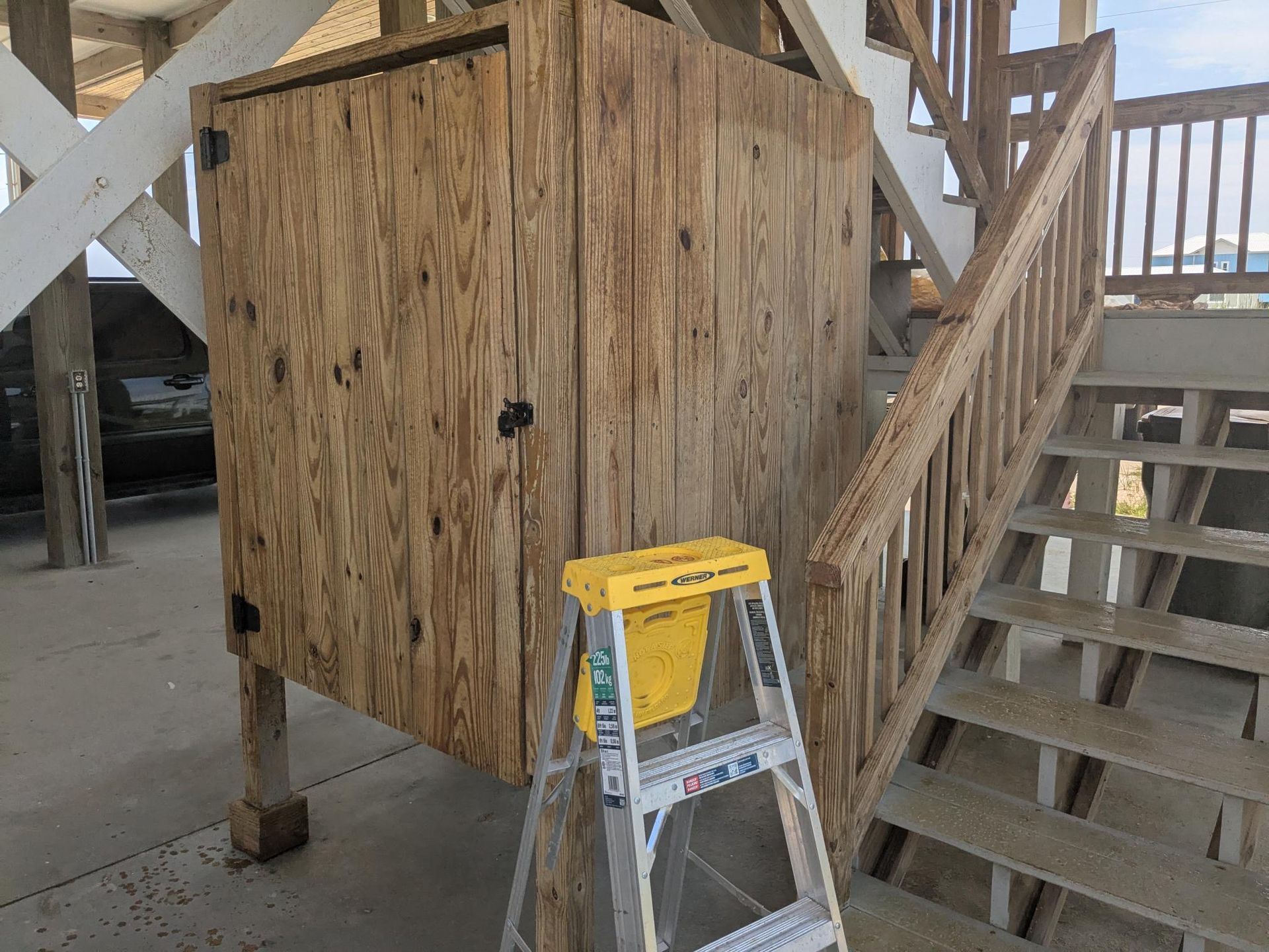 Outdoor wooden shower enclosure under a beach house. A stepladder is positioned in front.