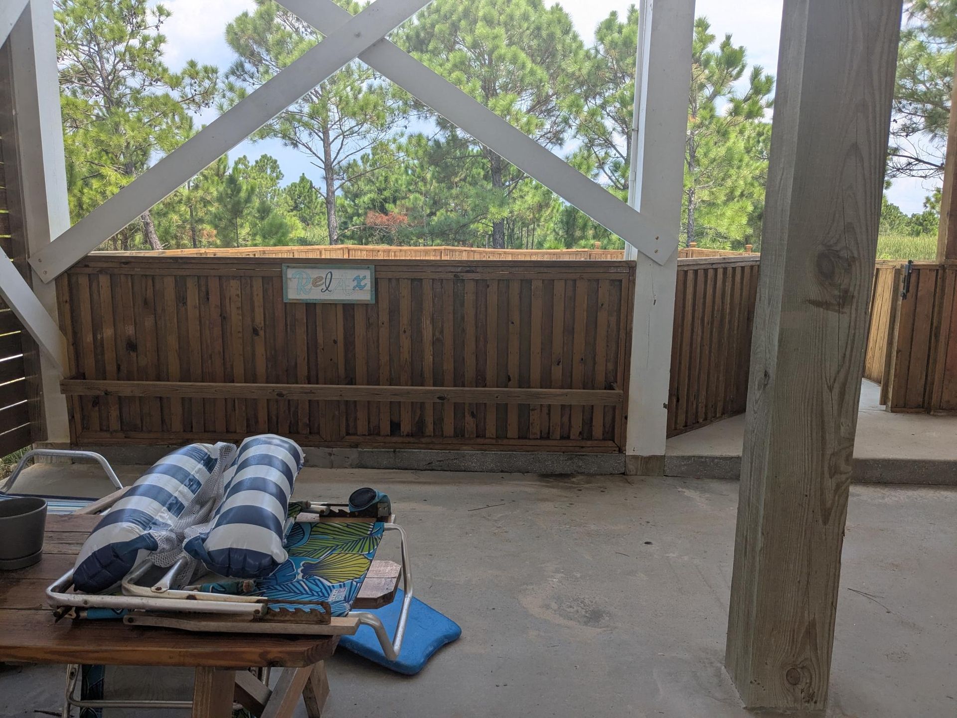 Covered patio with wooden walls, beams, and trees in the background. Lounge chairs and table in the foreground.