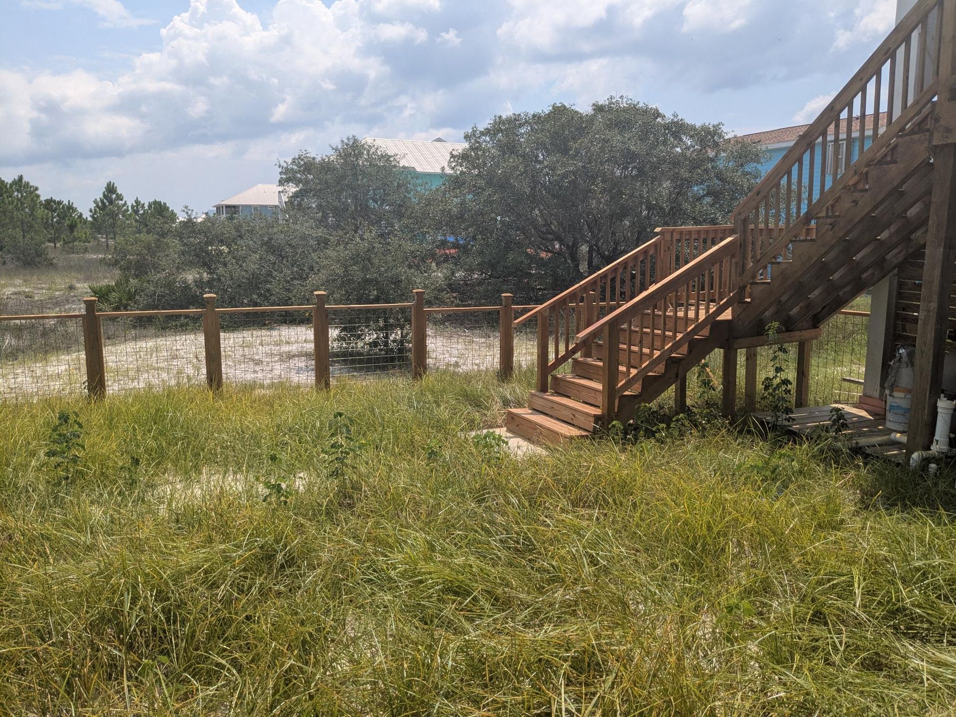 Wooden stairs leading up to a house with a surrounding wooden fence and tall grass.