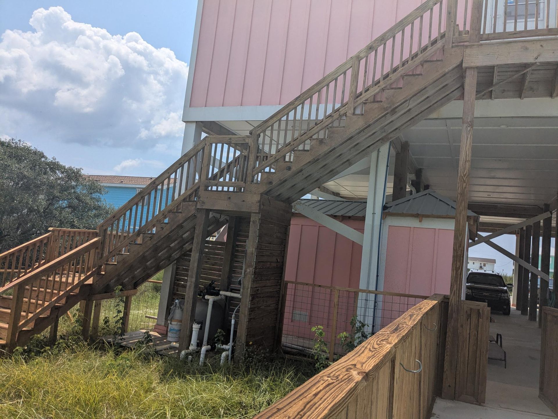 Wooden staircase leading up to a pink beach house on stilts with overgrown grass in the foreground.