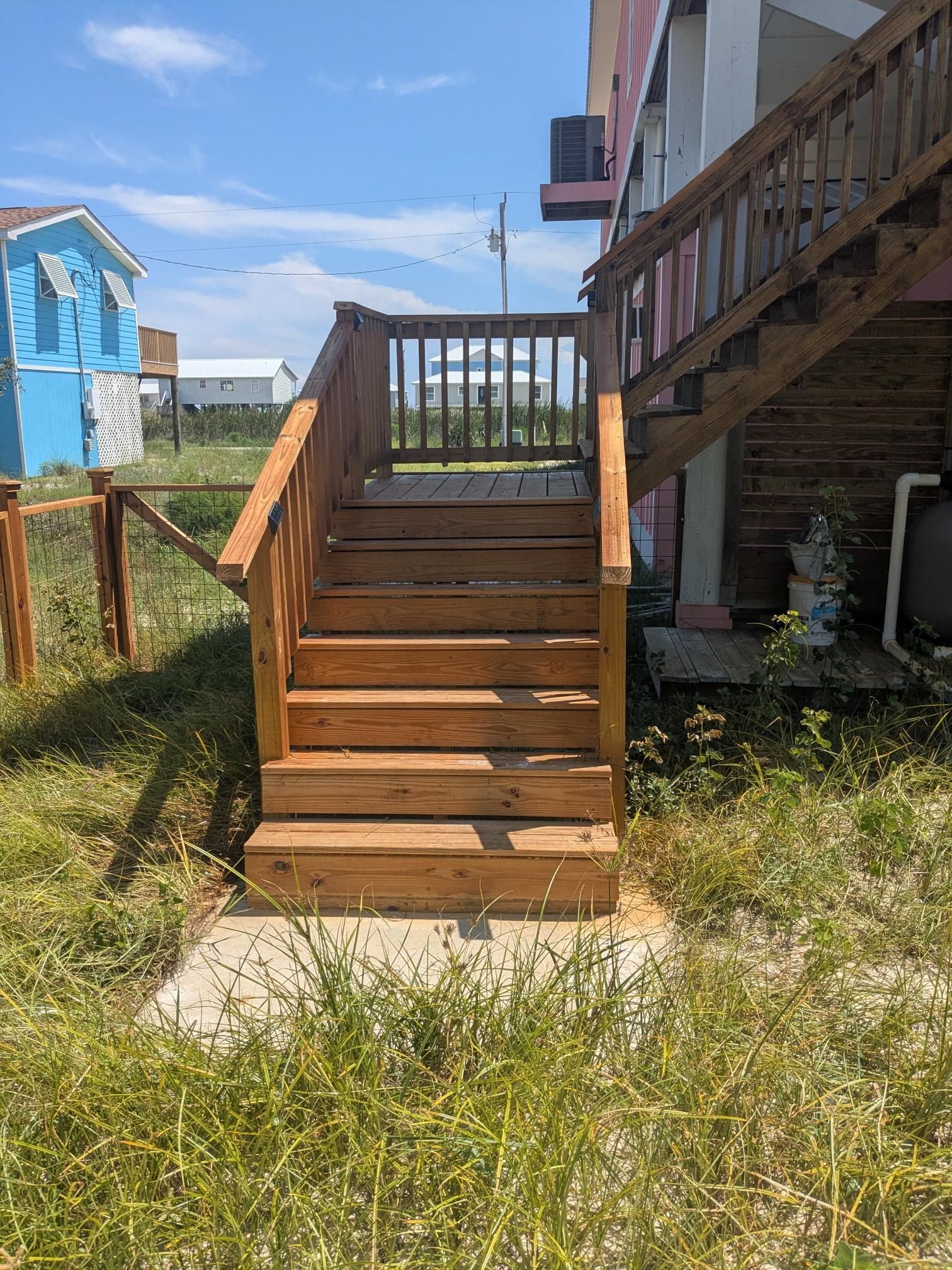 Wooden outdoor staircase leading up to a house; surrounded by grass and a blue sky.