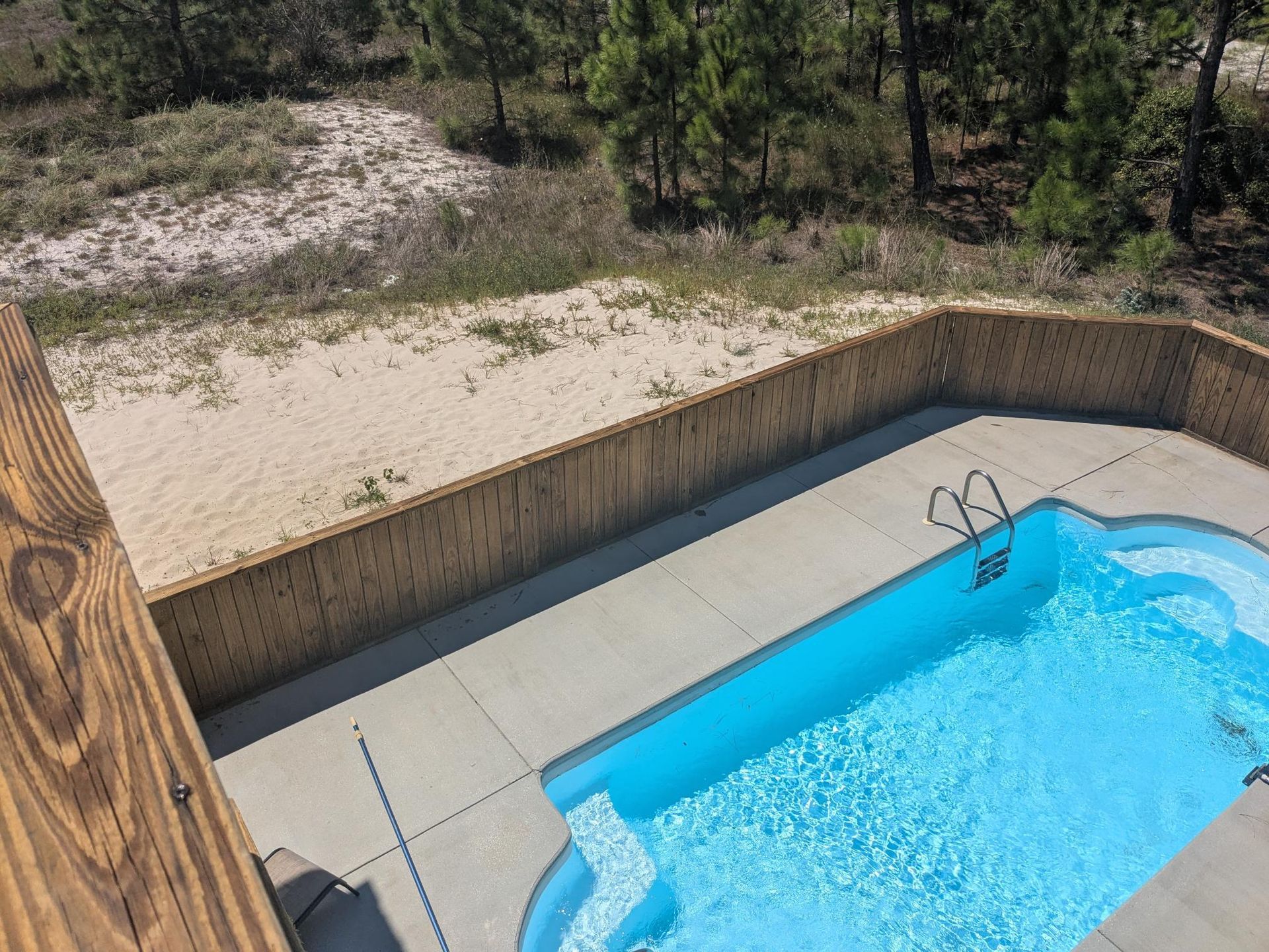 Swimming pool and surrounding concrete with wooden fence, next to a sandy dune and trees.