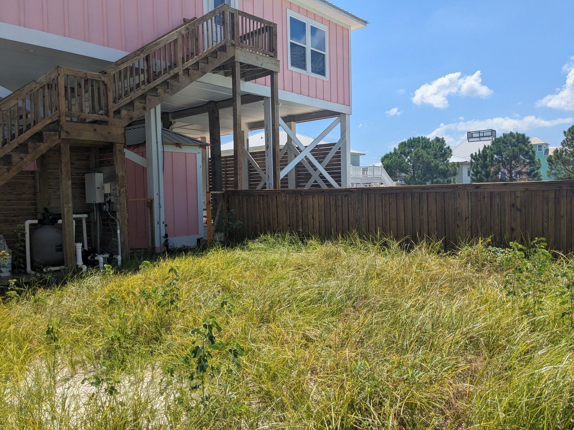 Pink house on stilts with wooden stairs, brown fence, overgrown grassy yard.