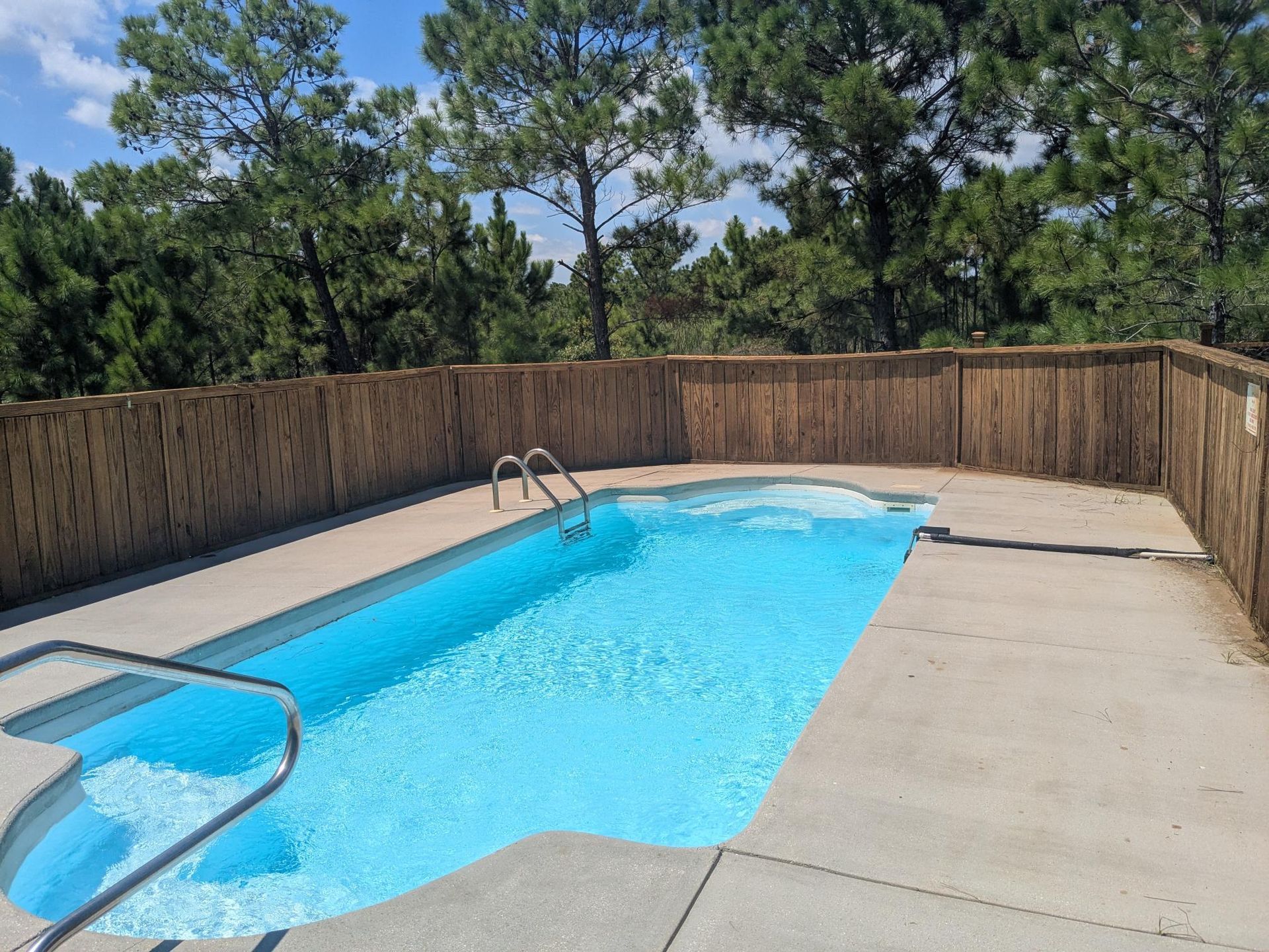 Swimming pool surrounded by concrete and a bamboo fence, with trees in the background.