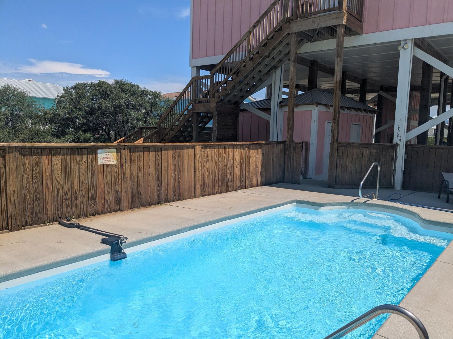 Swimming pool next to a wooden fence and pink stilt house with stairs on a sunny day.