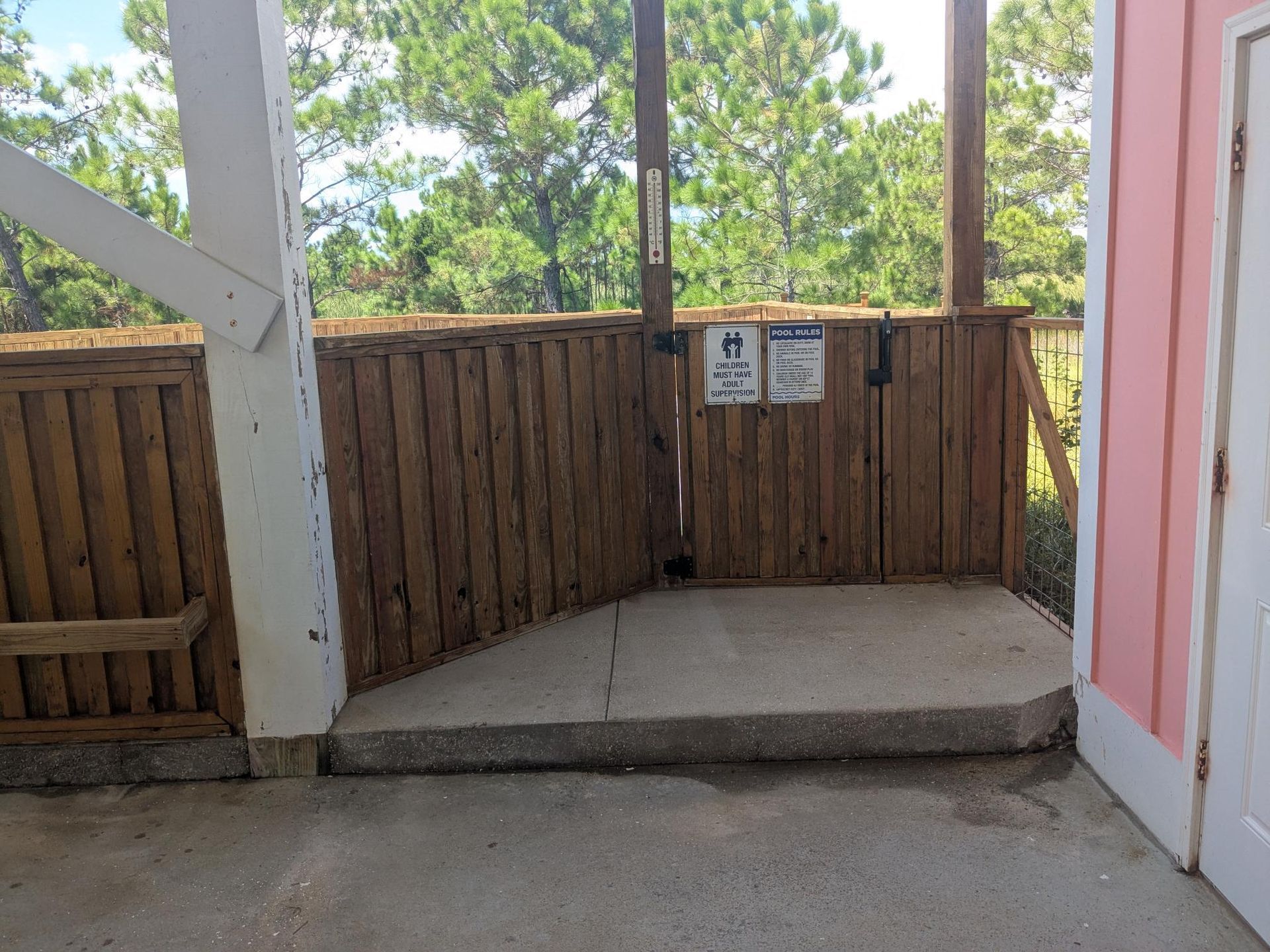 Wooden fence with gate, concrete platform, pink building side. Signage on gate. Trees in background.
