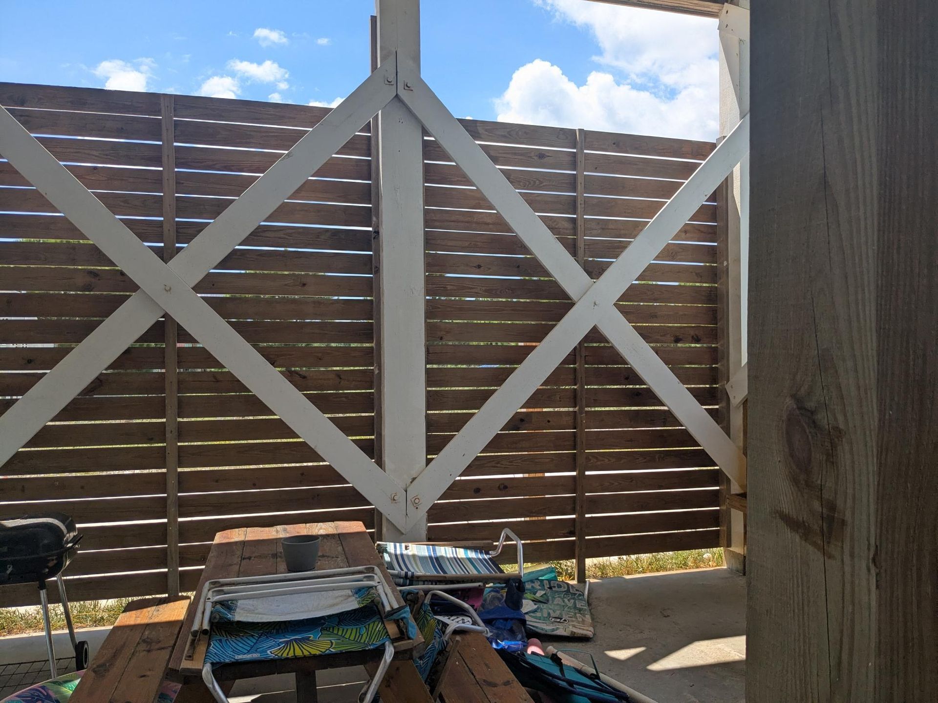 Wooden fence with white support beams, viewed from under a structure, with chairs and picnic table visible.