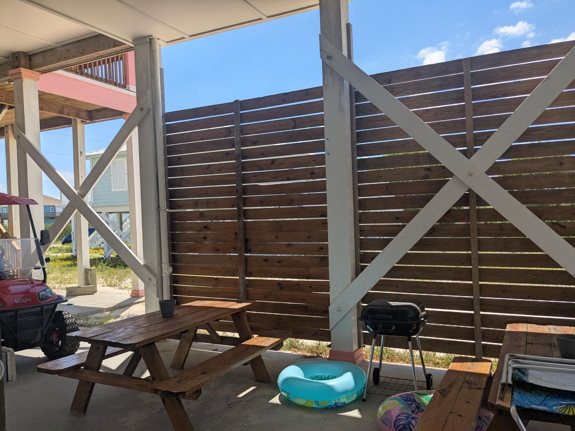 Covered patio with picnic table, grill, and beach float; horizontal wooden fence and support beams.