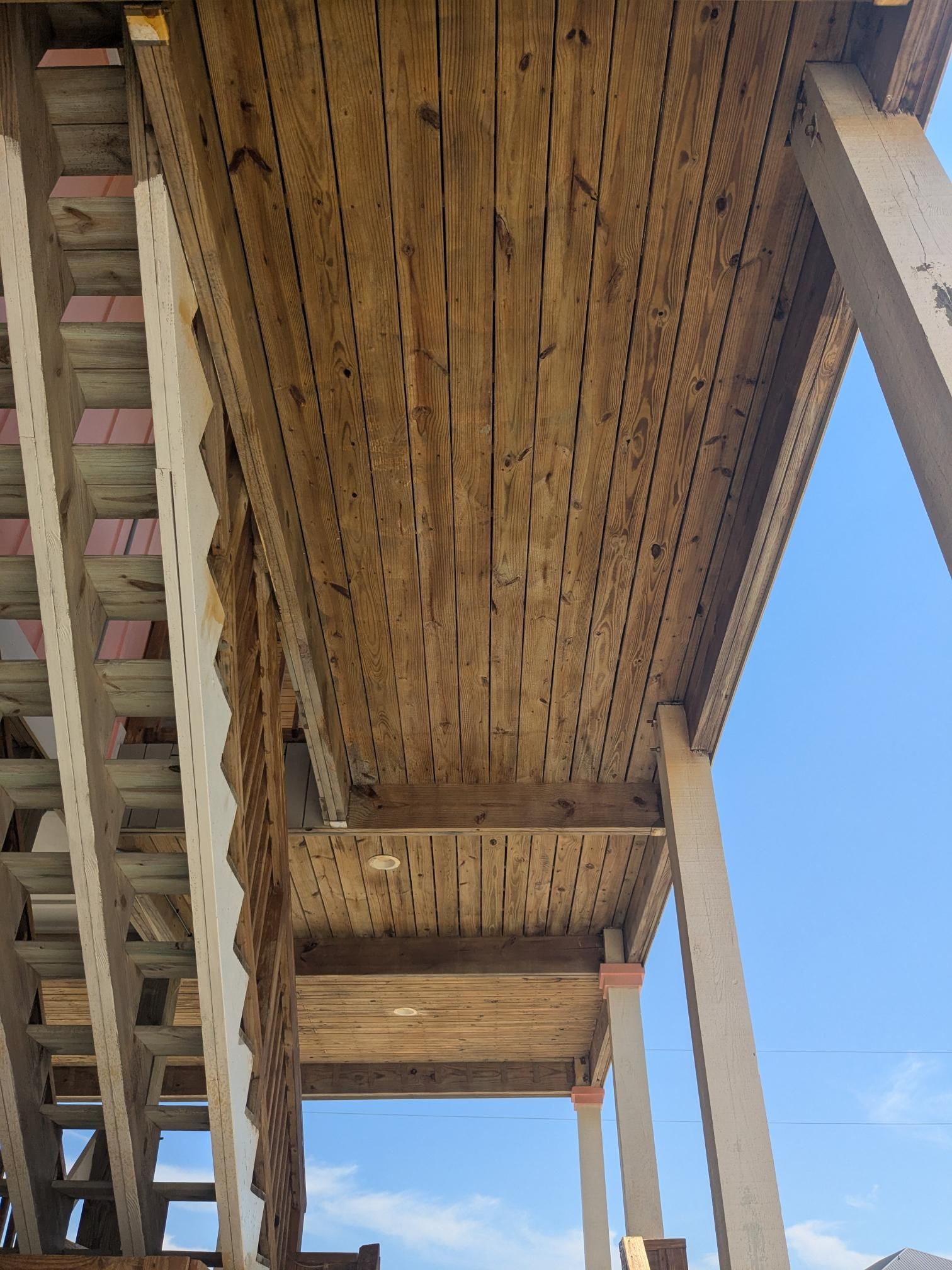 Underside of a wooden porch with support beams, weathered boards, and visible nails. Bright sky background.