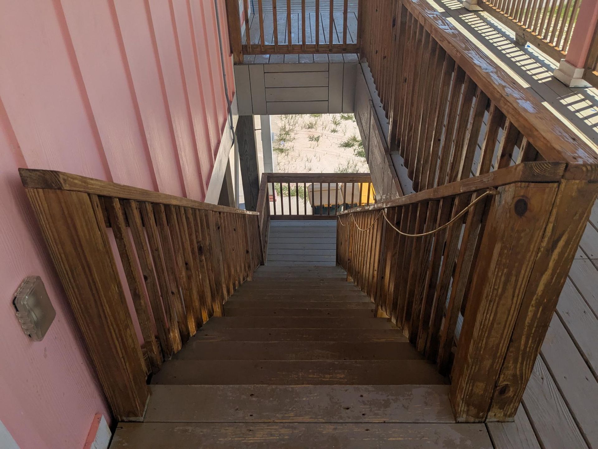 Wooden outdoor staircase leading downward, with brown railings and a pink wall on the left.