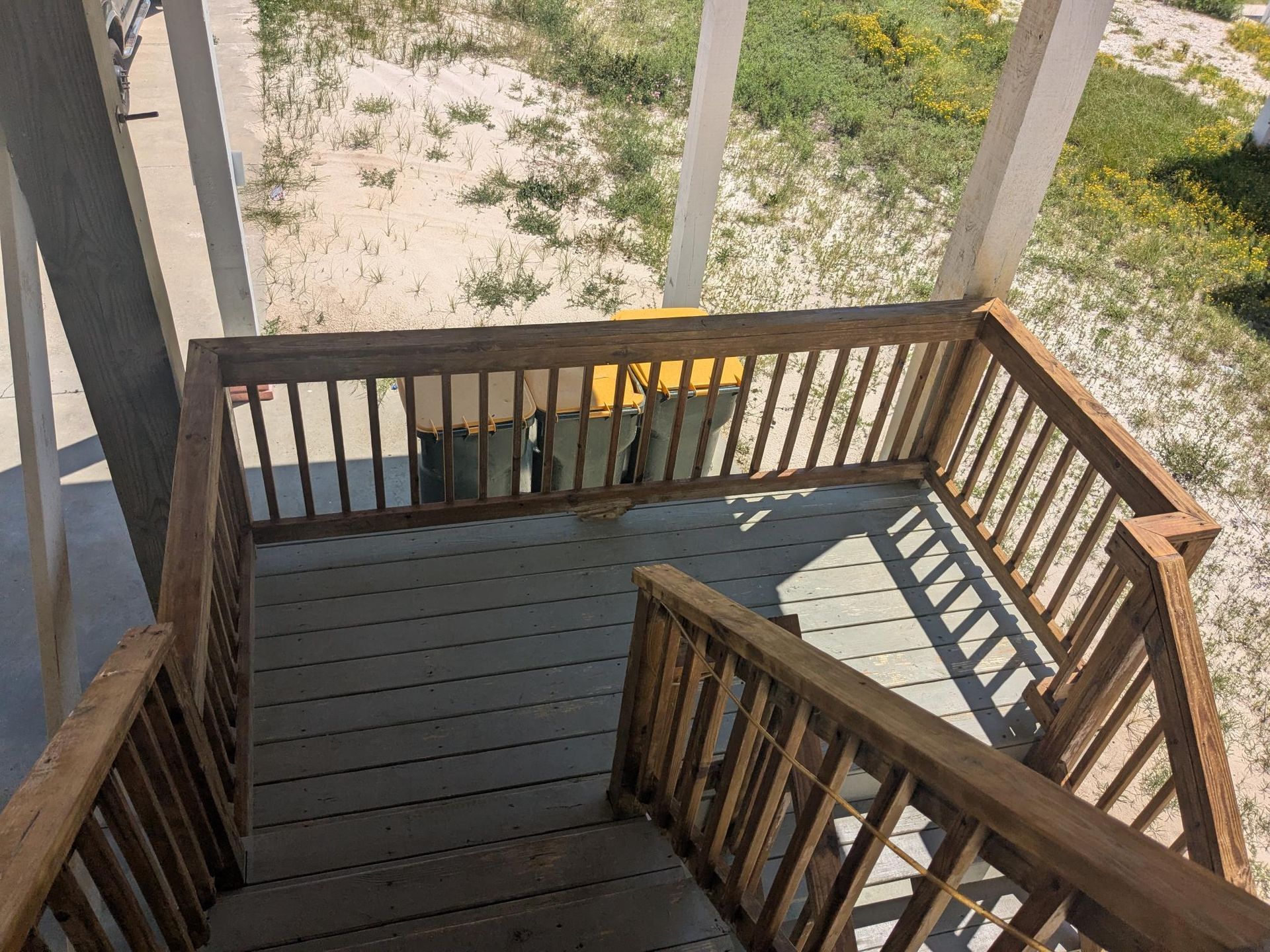 Wooden deck and stairs with railing. Sunlit outdoor area with sand and grass visible.