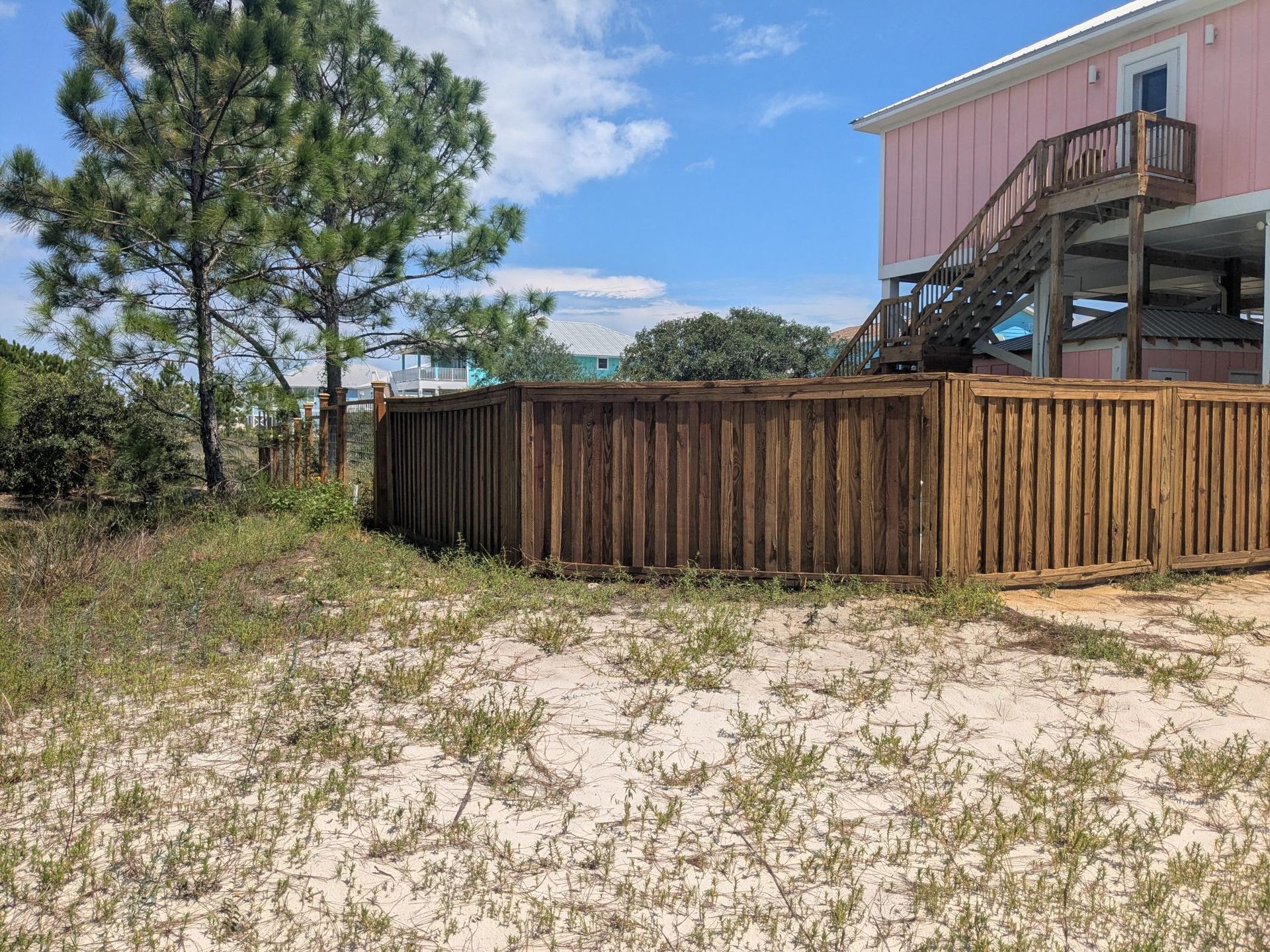 Wooden fence surrounds a beach house. The sky is blue. Sand and sparse vegetation are in the foreground.
