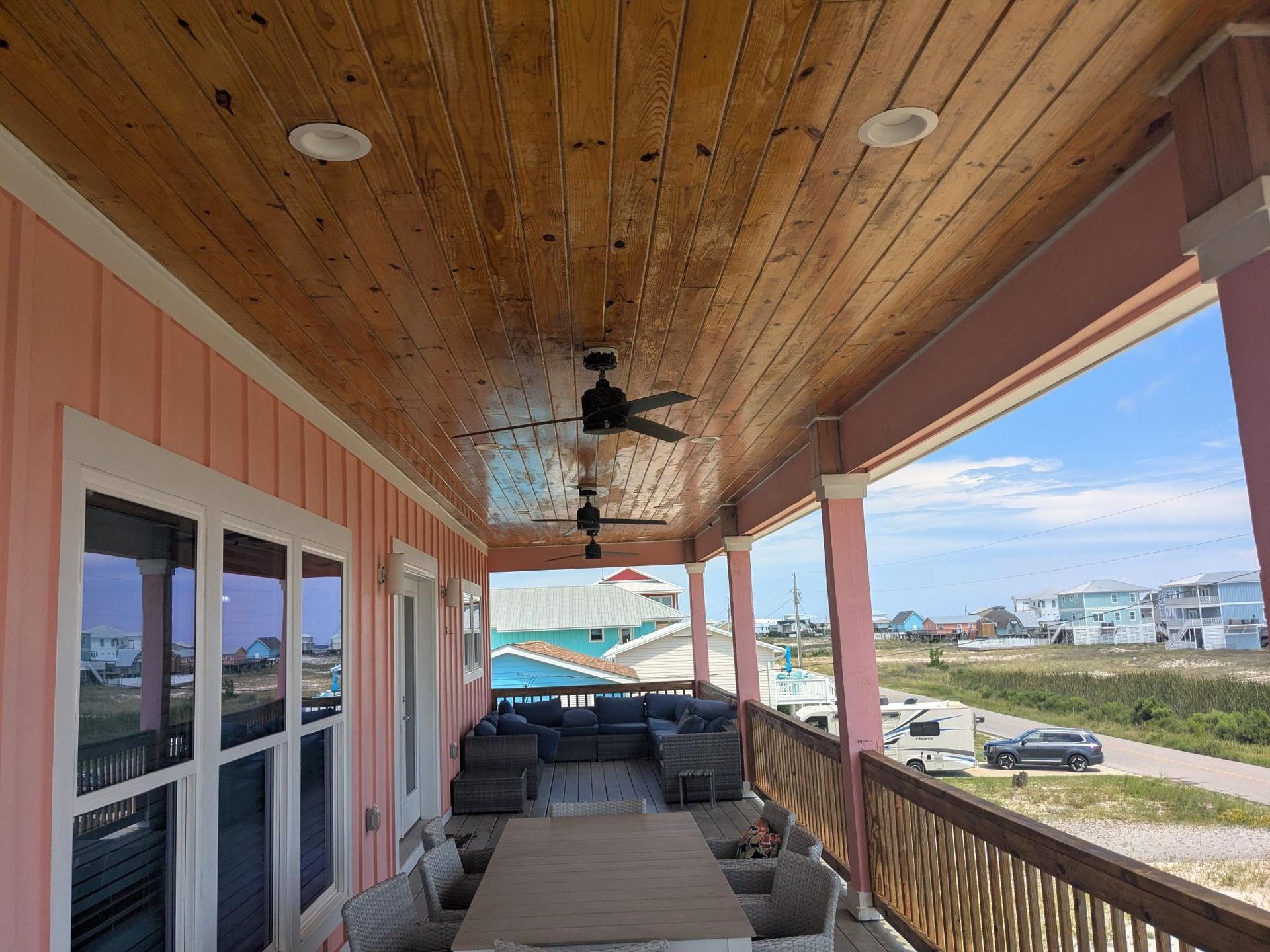 Covered outdoor porch with pink walls, wood ceiling, and ceiling fans, overlooking a sunny coastal landscape.