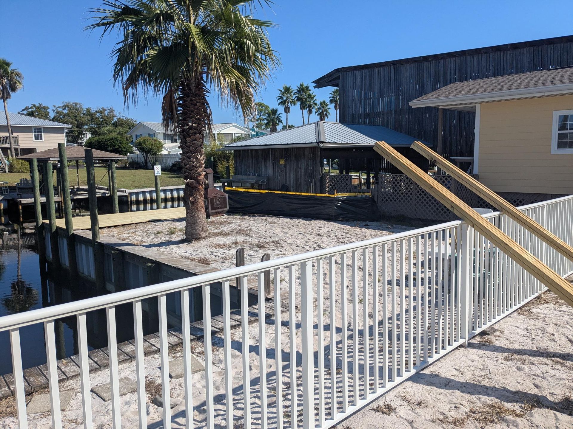 White fence frames a waterfront yard with a dock, palm tree, and buildings.
