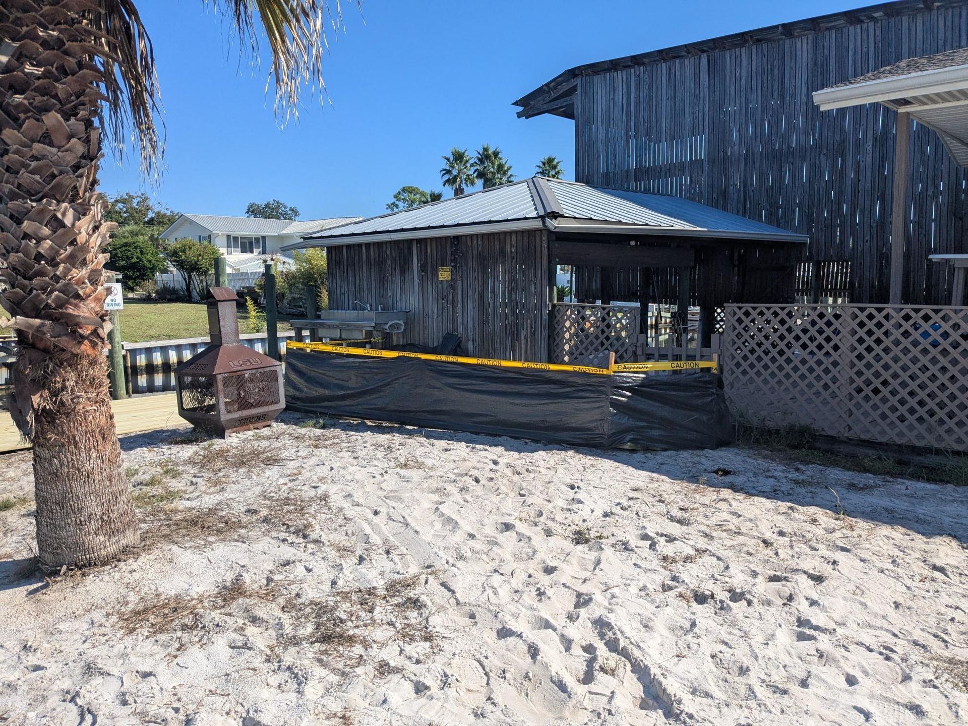Beachfront wooden structure, fenced off with caution tape. Sand foreground, palm tree on left.
