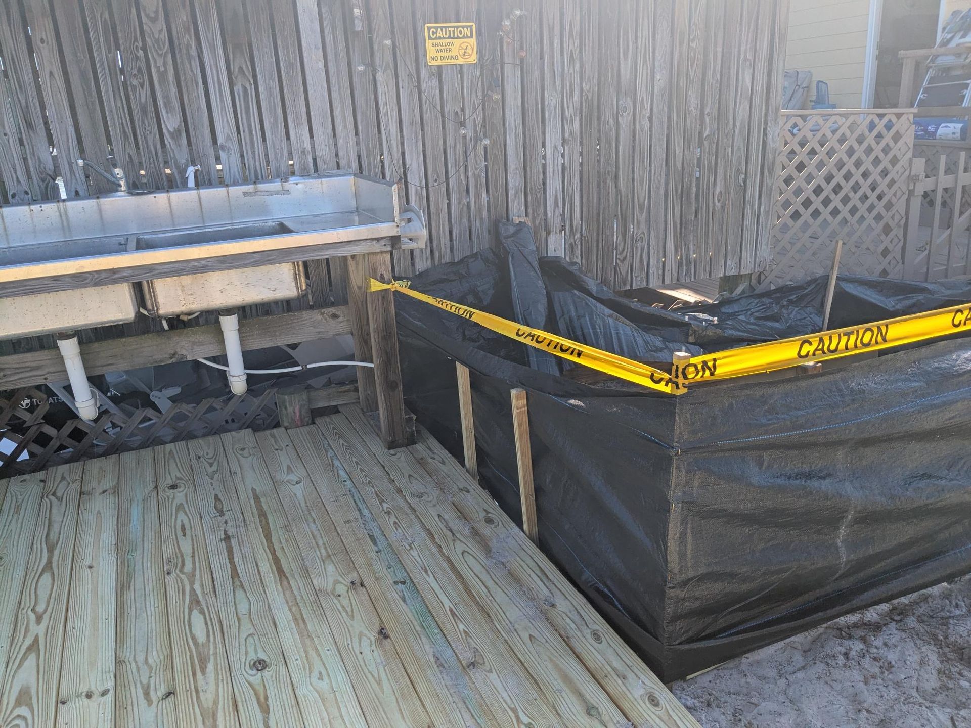 Outdoor scene with a wooden deck, a sink, and a black tarp-lined area marked with caution tape.