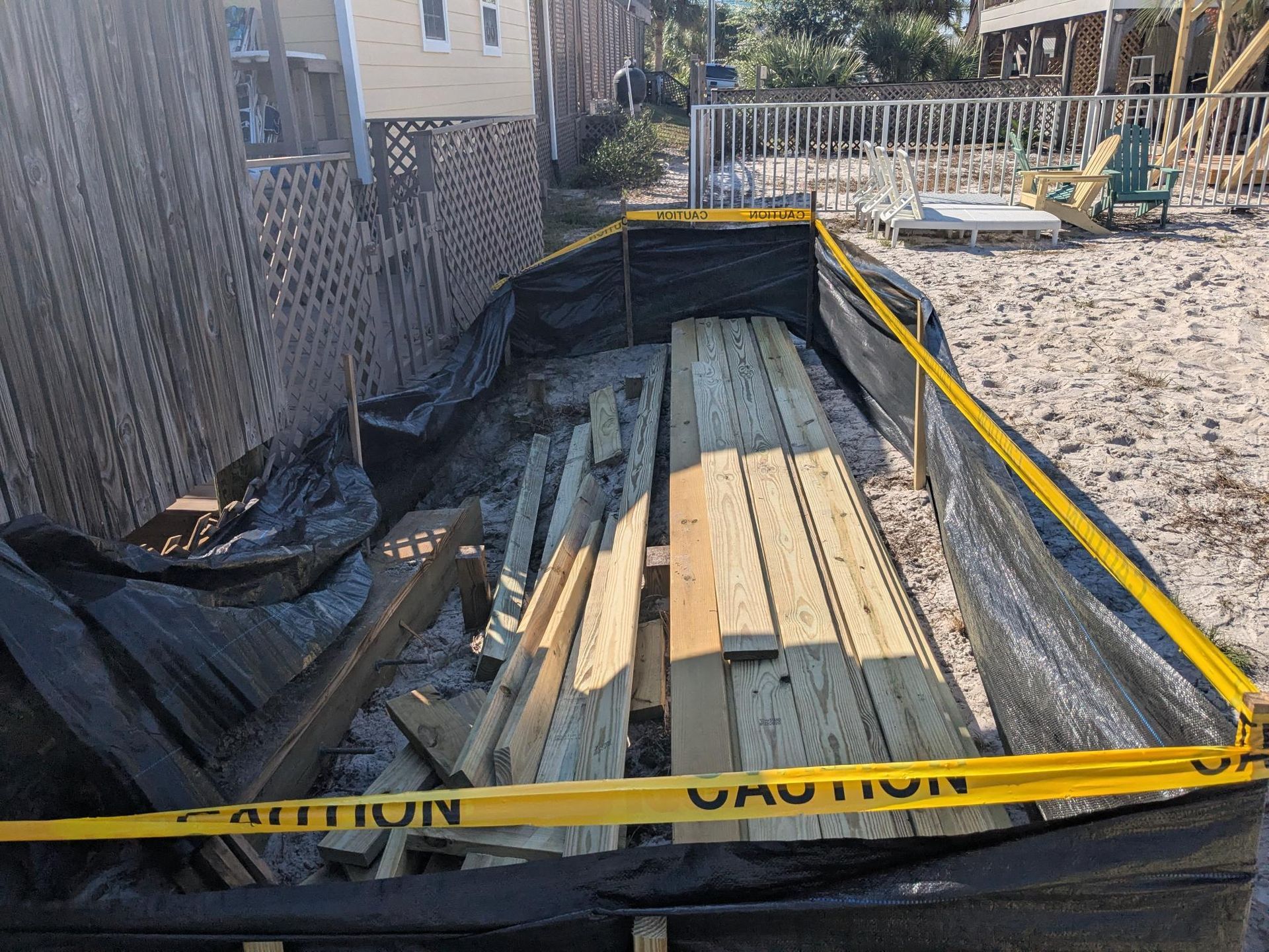 Construction debris, including wooden planks, contained by black barrier with caution tape. Near a building and beach.
