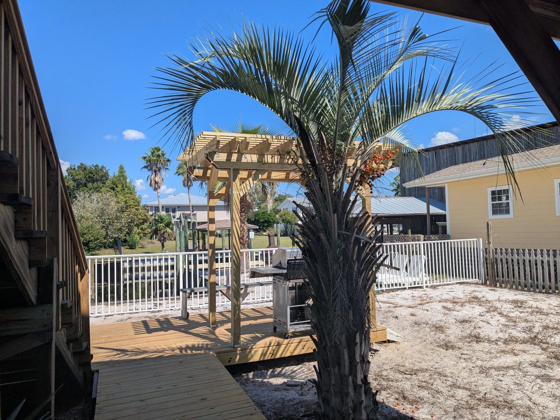 Wooden deck with pergola and palm tree. Yellow building and white fence in background.