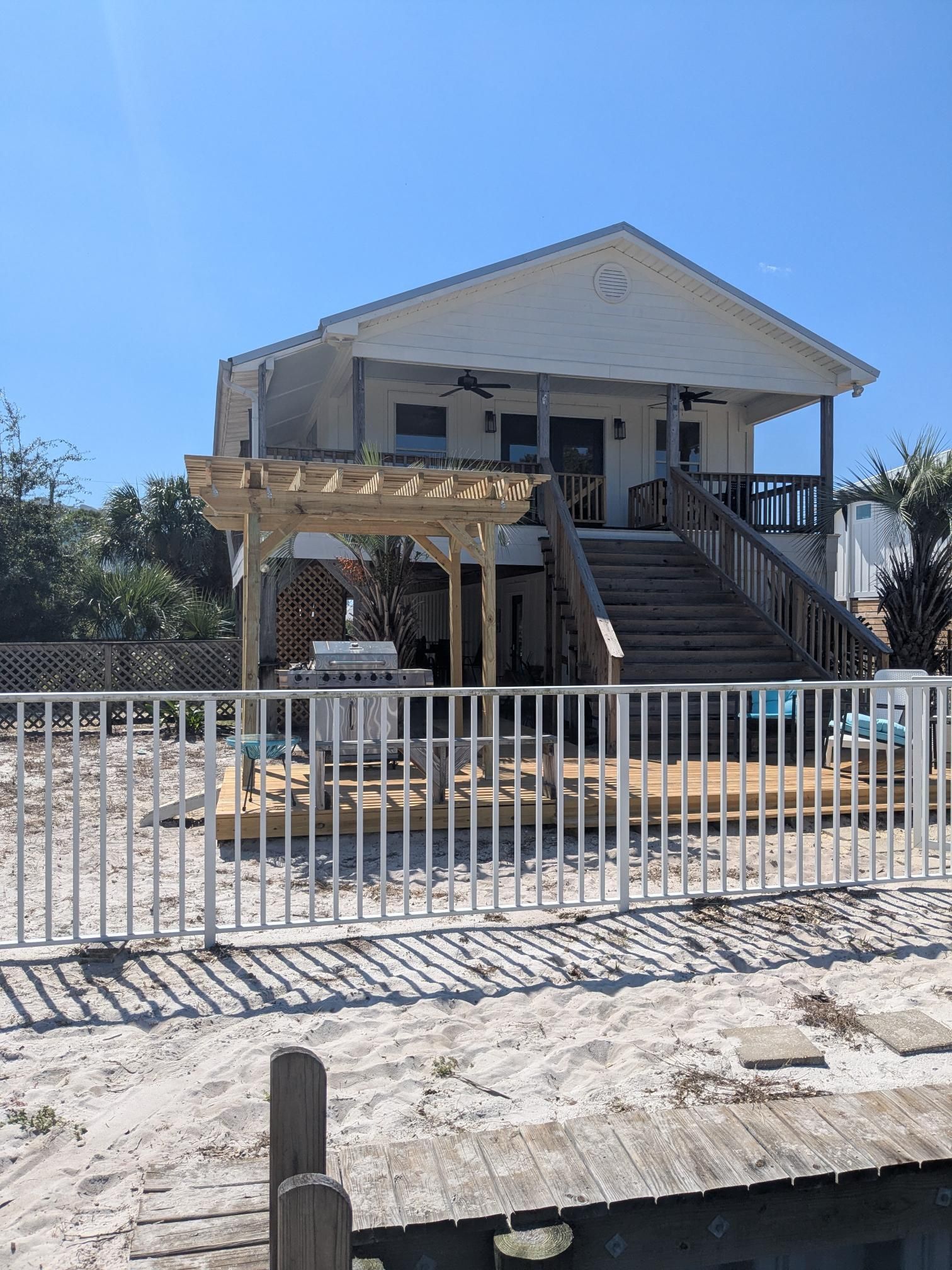 Beach house with pergola, deck, and stairs. White siding, wooden railing and deck, blue sky.