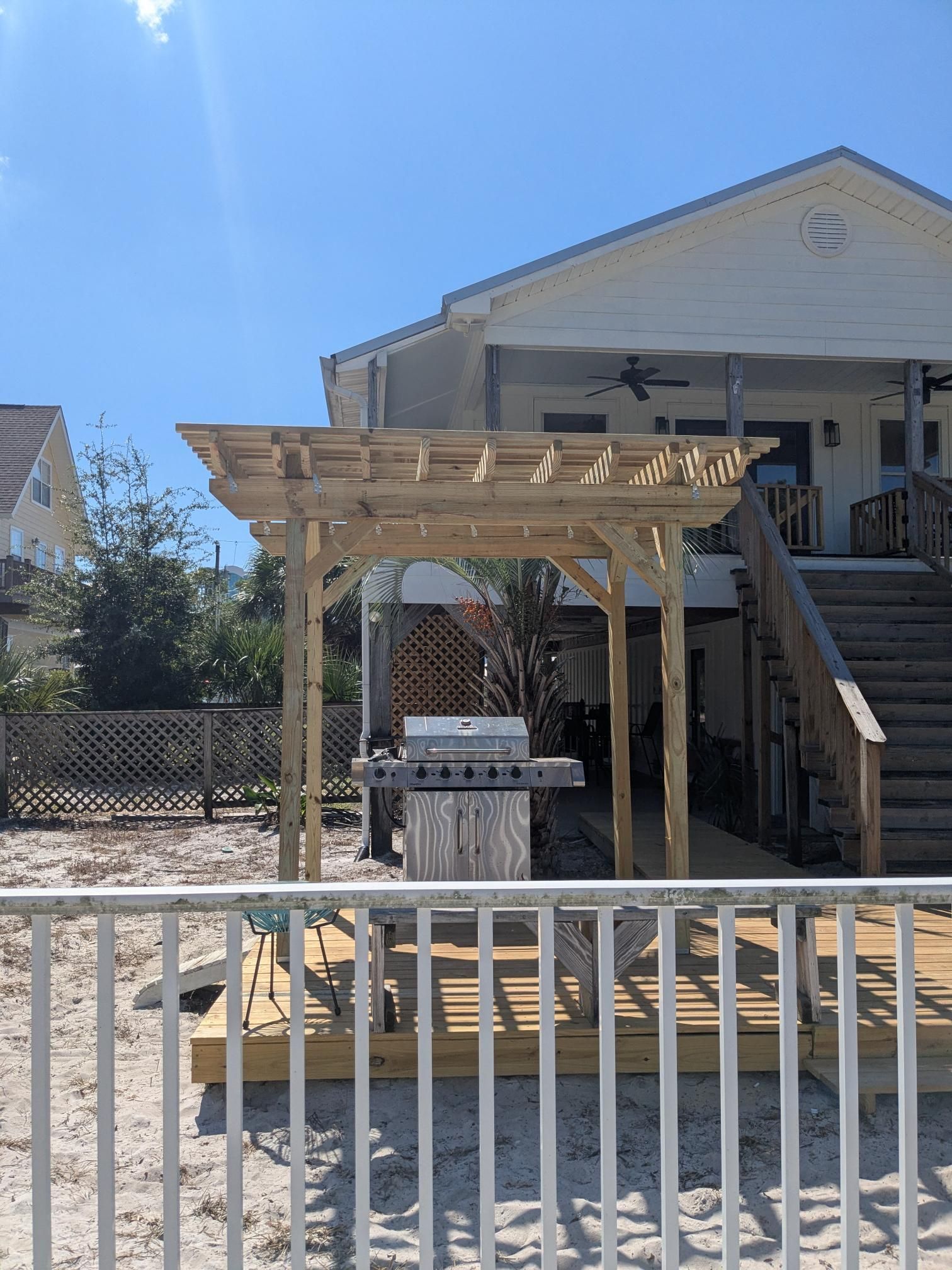 Wooden pergola over an outdoor grill on a wooden deck. A two-story house is in the background. Sunny day.