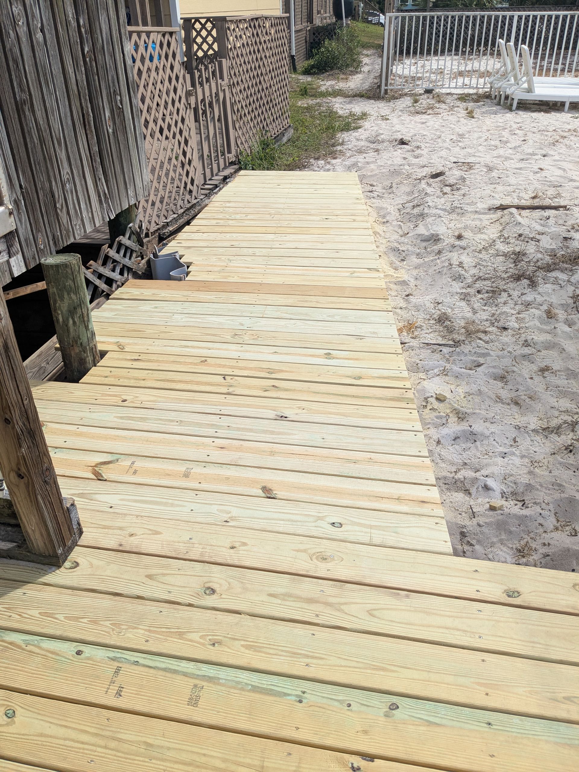 Wooden boardwalk leading to sandy beach; structure on left, fence and white railing in background.