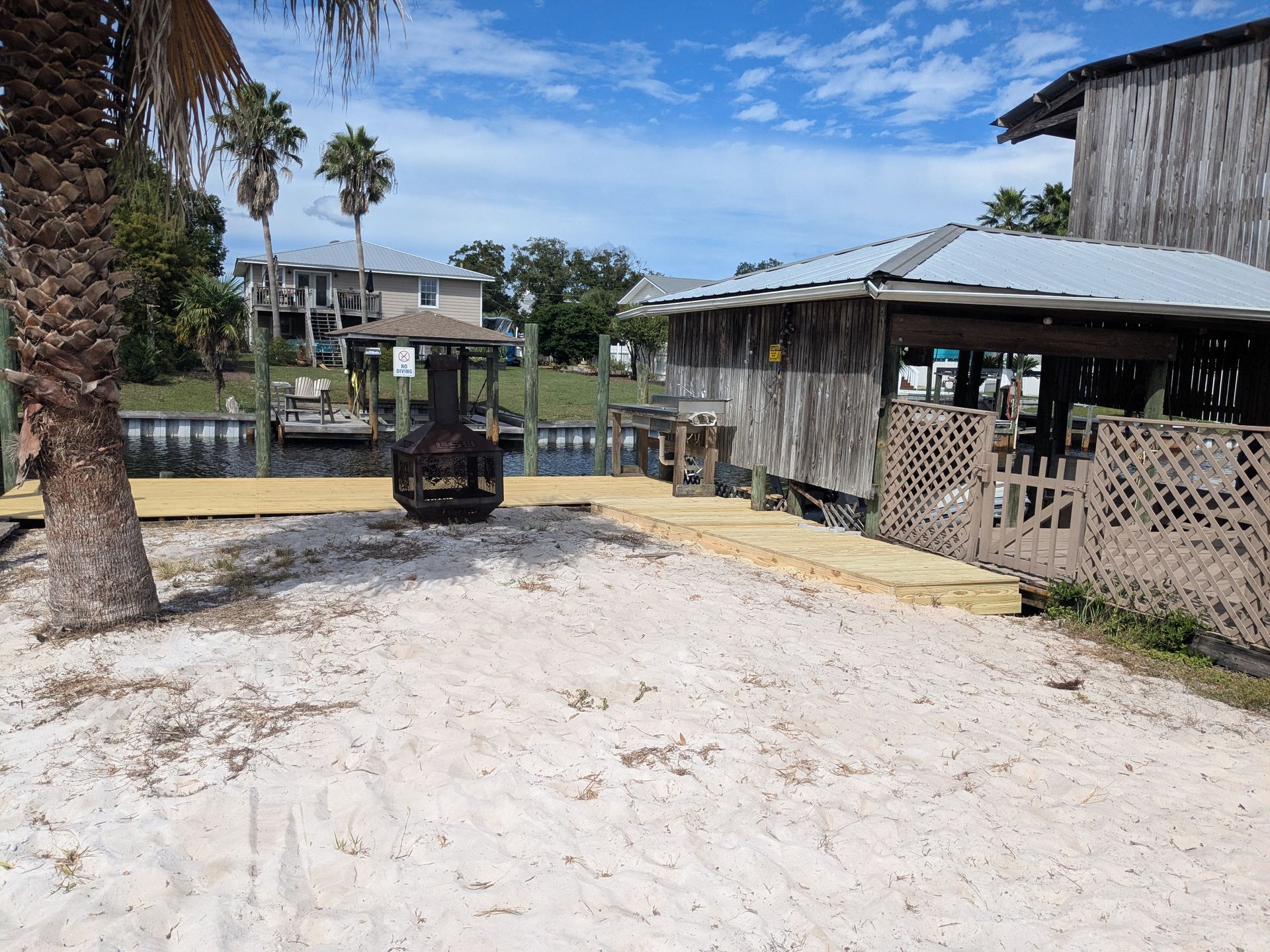 Sandy beach with wooden deck, weathered building, and fire pit. Water and homes visible in the background.
