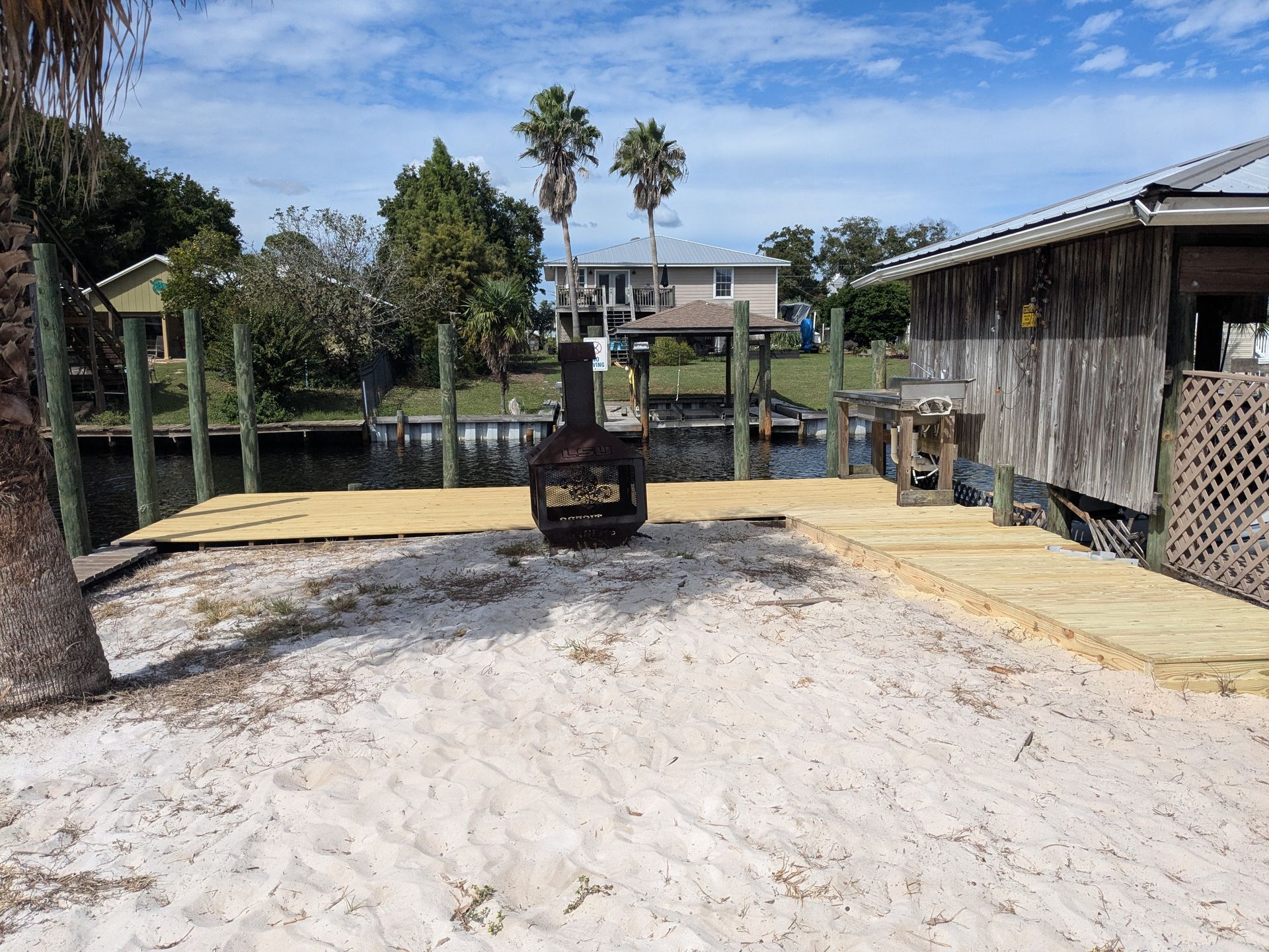 Sandy waterfront with wooden dock, fire pit, canal and house in background.