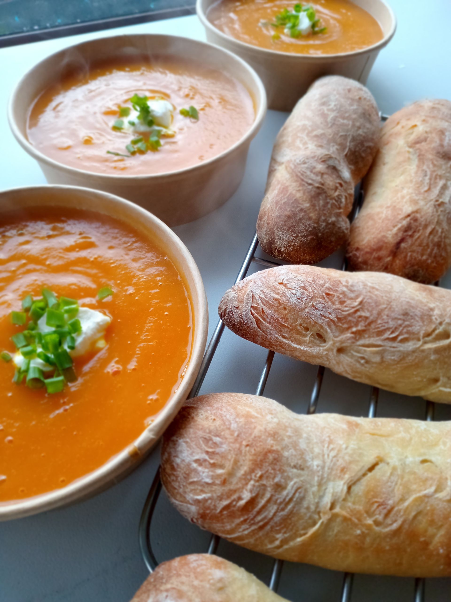 Bowls of orange soup with bread rolls, garnished with cream and chives, on a table.