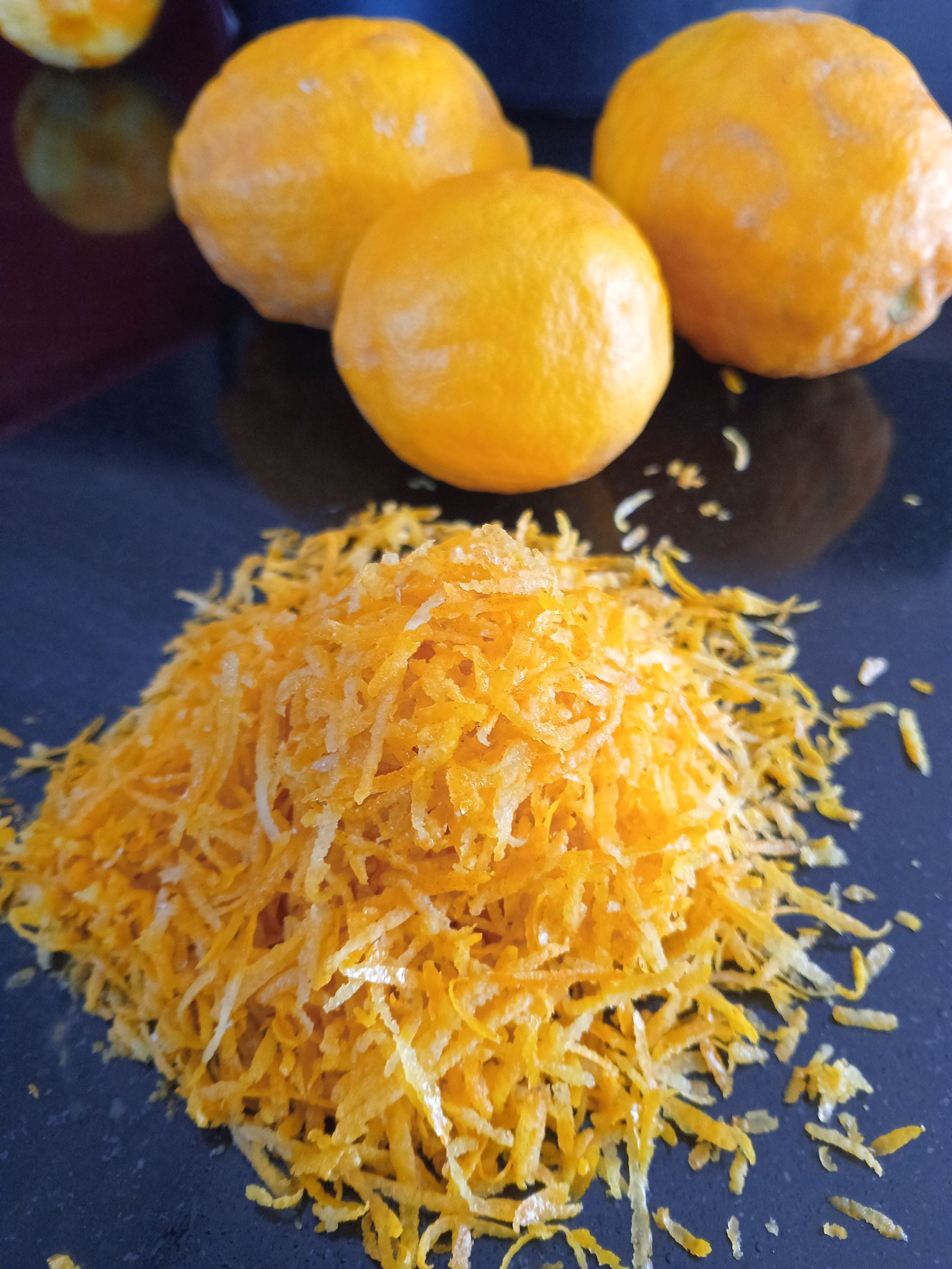 Pile of bright orange citrus zest with whole oranges in the background on a dark surface.
